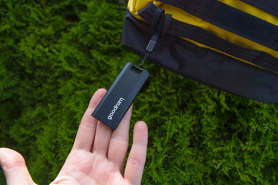 Dłoń trzyma czarny pendrive Goodram. Żółto-czarna torba jest w tle, a zielone liście wypełniają scenę.