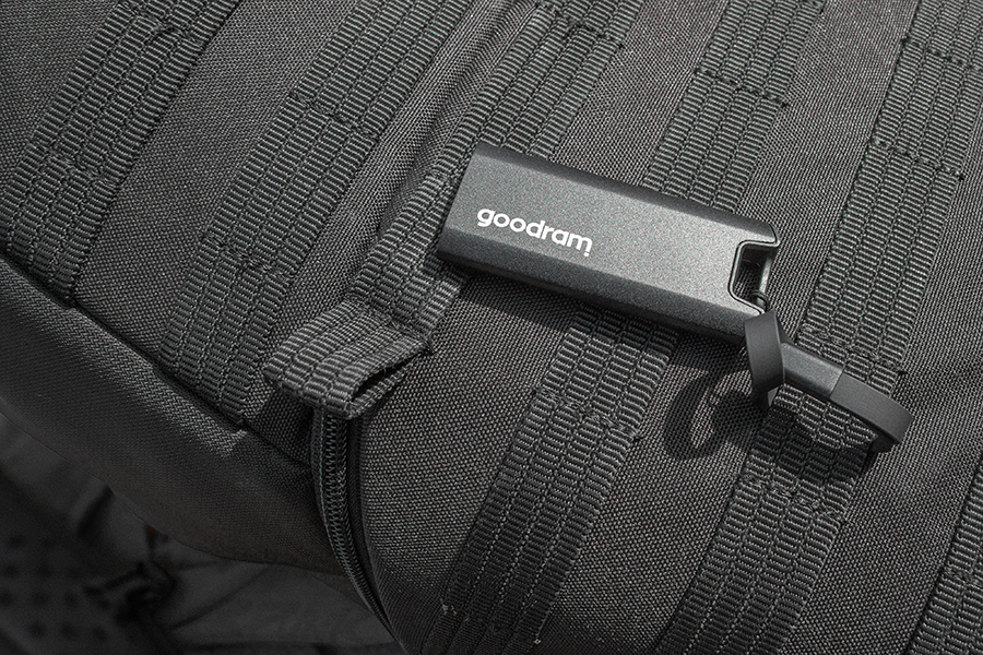 Czarny pendrive z napisem 'goodram' leży na czarnym plecaku. Plecak ma teksturowaną powierzchnię z wszytymi paskami.