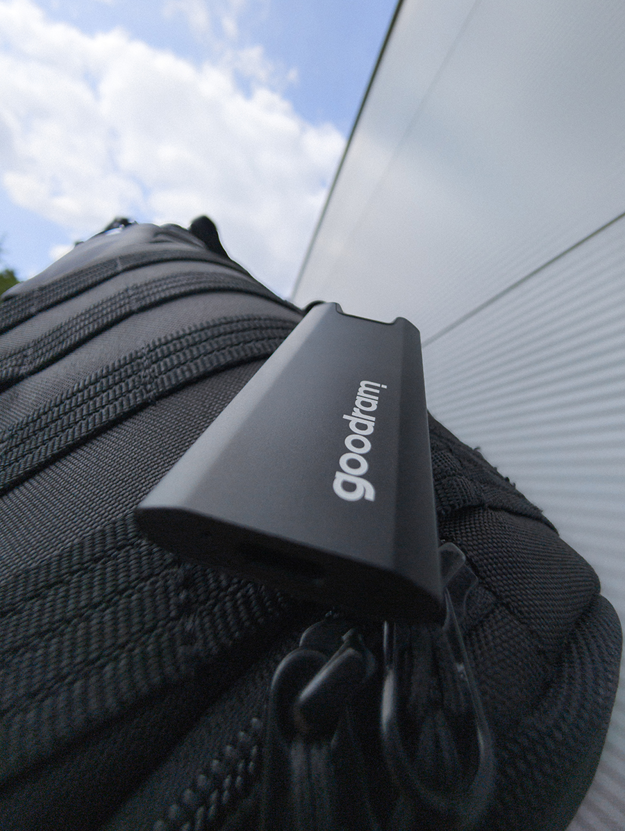 Czarny pendrive Goodram spoczywa na czarnej, teksturowanej powierzchni, prawdopodobnie torbie. Biały budynek i błękitne niebo w tle.