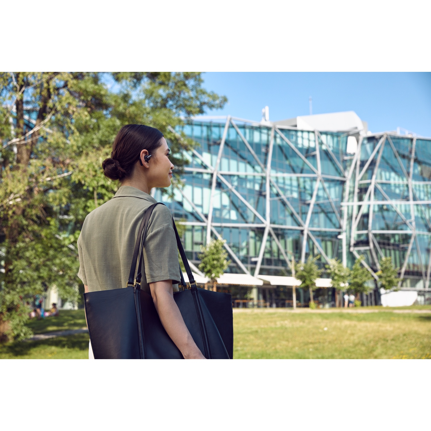 Une femme avec des écouteurs et un sac marche devant un bâtiment moderne en verre. Des arbres et de l'herbe sont au premier plan.