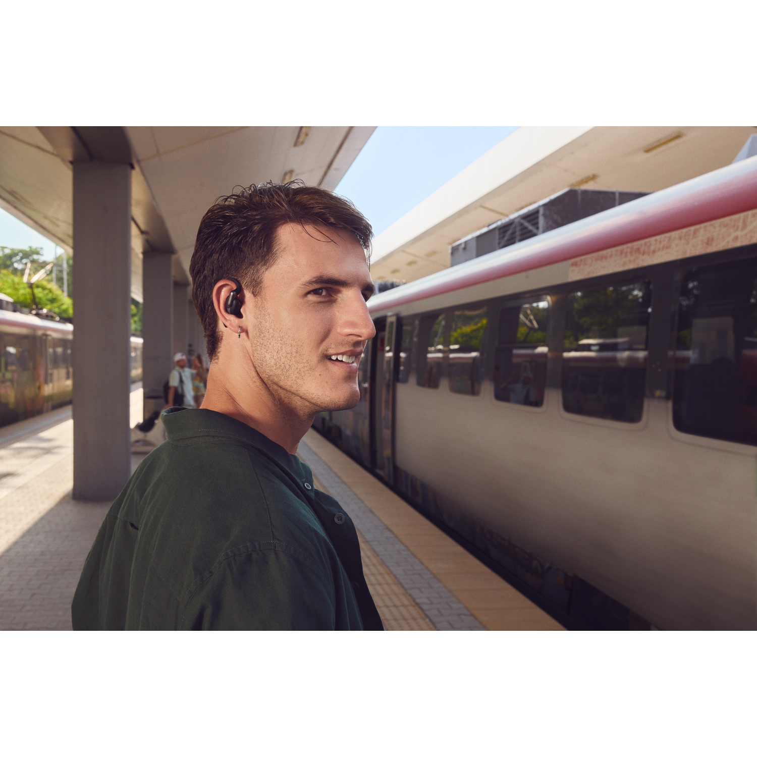 Un homme avec un écouteur se tient sur un quai de gare. Un train passe. Il porte une chemise verte et sourit.