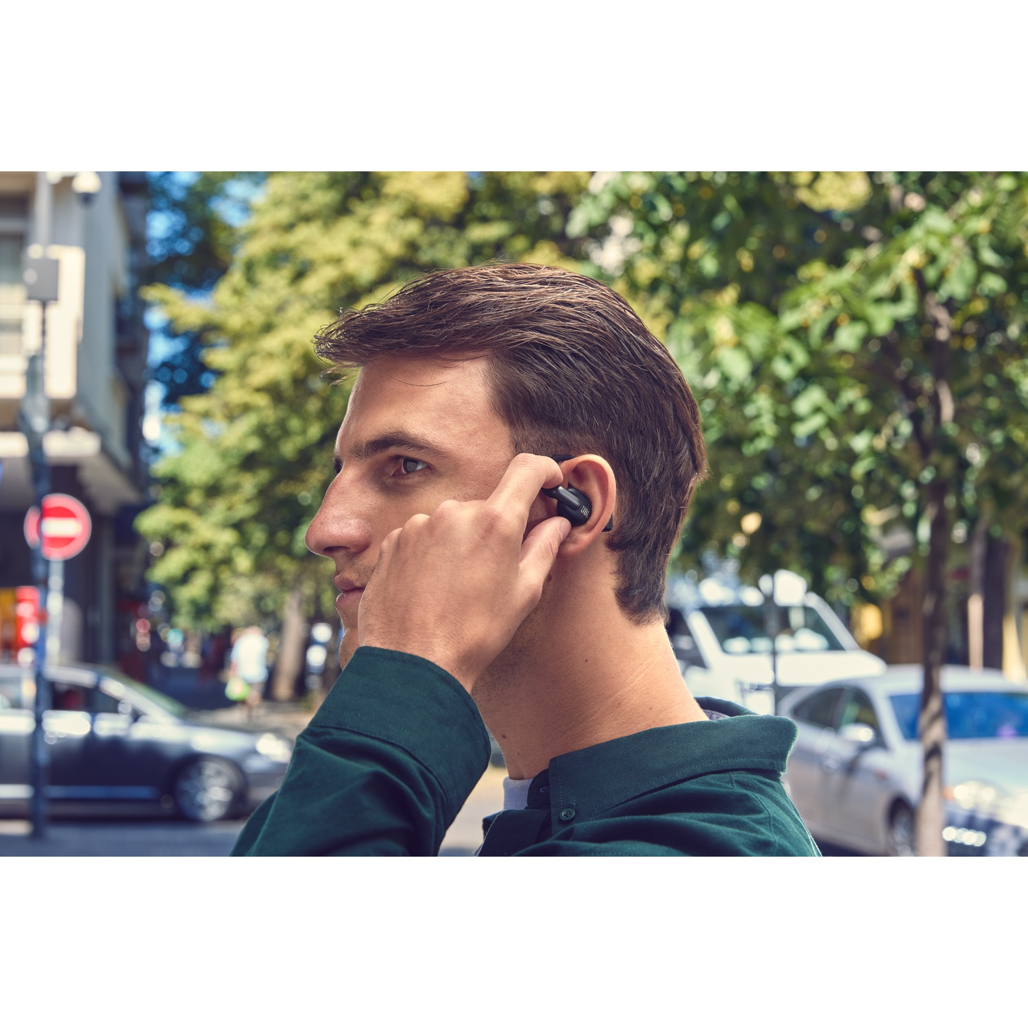 Un homme en veste verte met un écouteur noir dans son oreille. Voitures et arbres en arrière-plan.