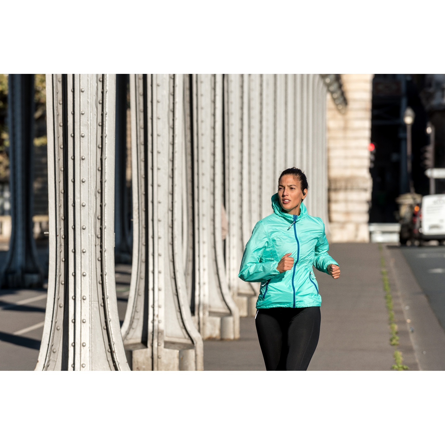 Femme courant sur un pont. Elle porte une veste turquoise et un legging noir. Colonnes métalliques à gauche. Voitures en arrière-plan.