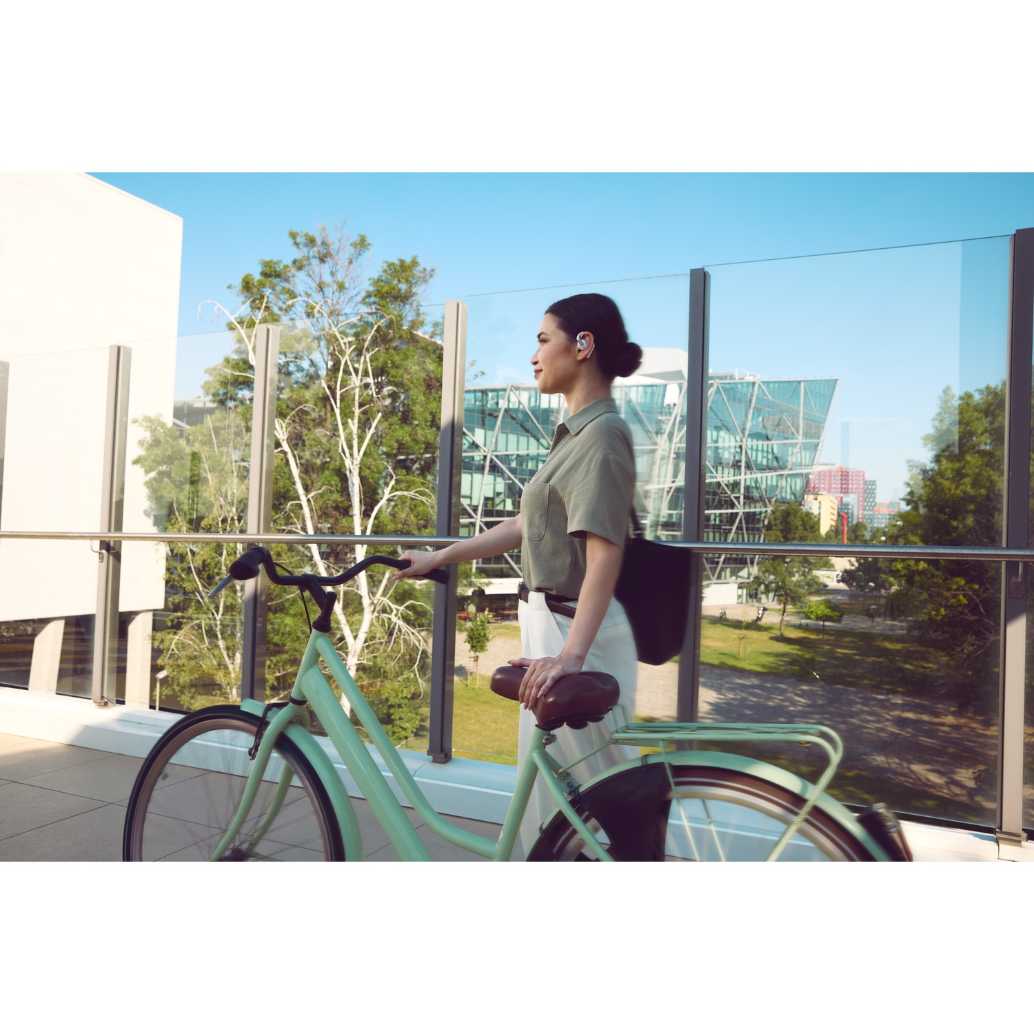Femme avec un vélo, marchant sur un pont. Bâtiments et arbres en arrière-plan. Le vélo est vert menthe.