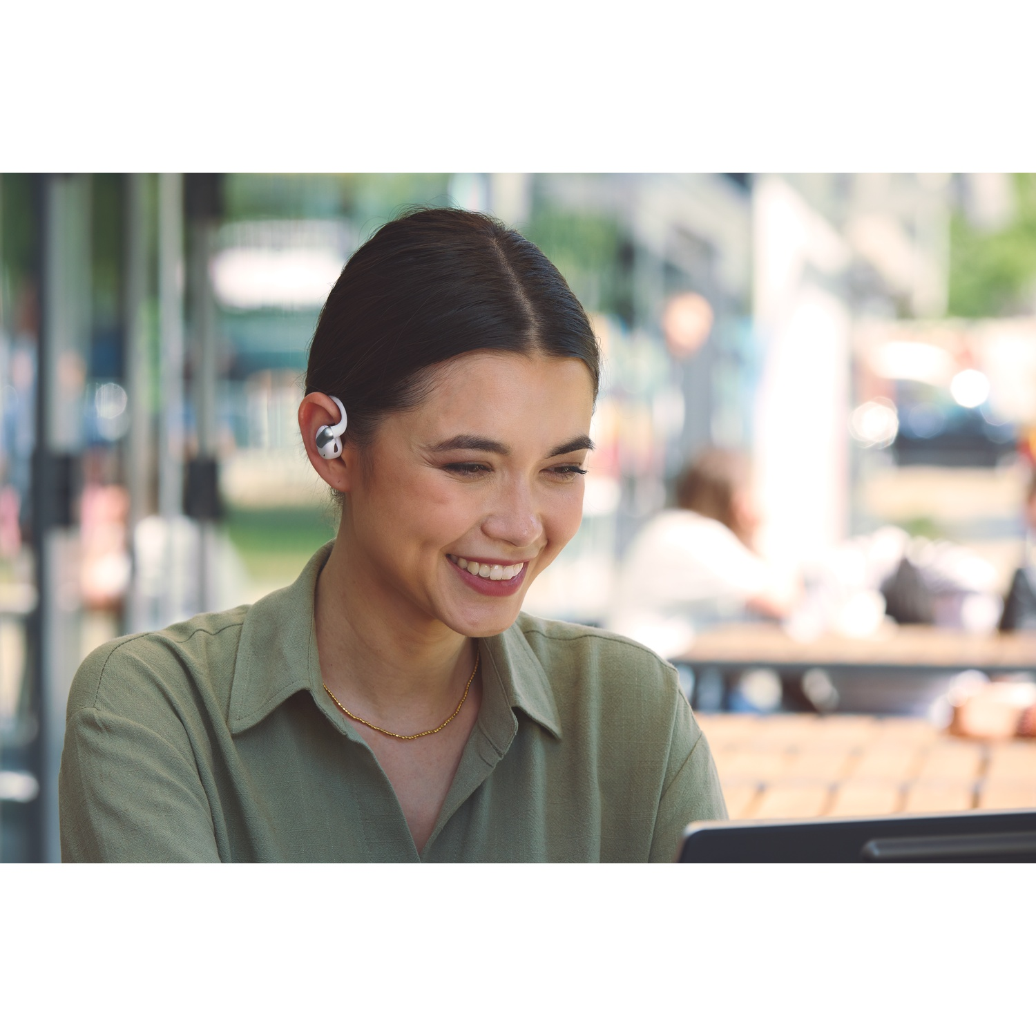 Une femme souriante portant des écouteurs et une chemise verte regarde un ordinateur portable. Elle a un collier en or. Le fond est flou.