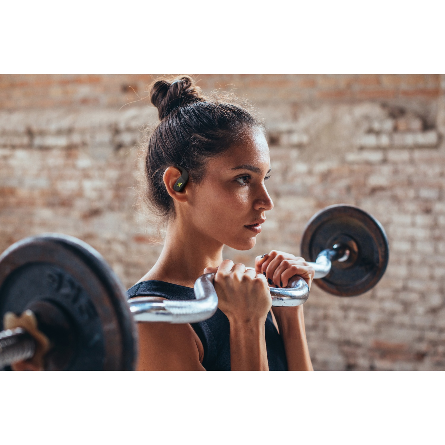 Femme dans une salle de sport avec haltère, portant des écouteurs. Fond de mur en briques. Expression concentrée en soulevant des poids.