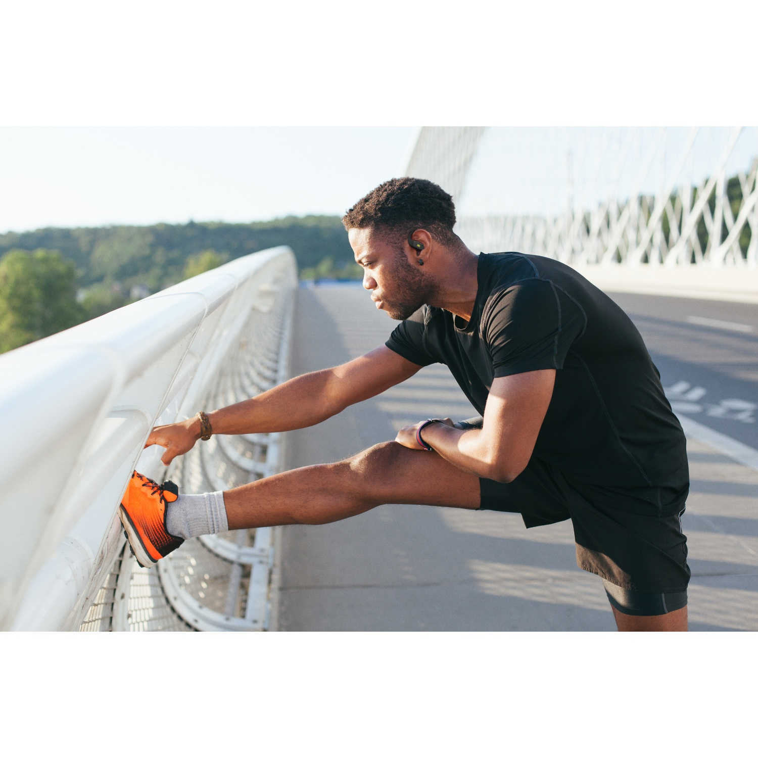 Un homme noir en tenue de sport s'étire la jambe sur un pont, tenant une rampe blanche. On aperçoit des baskets orange et noires.