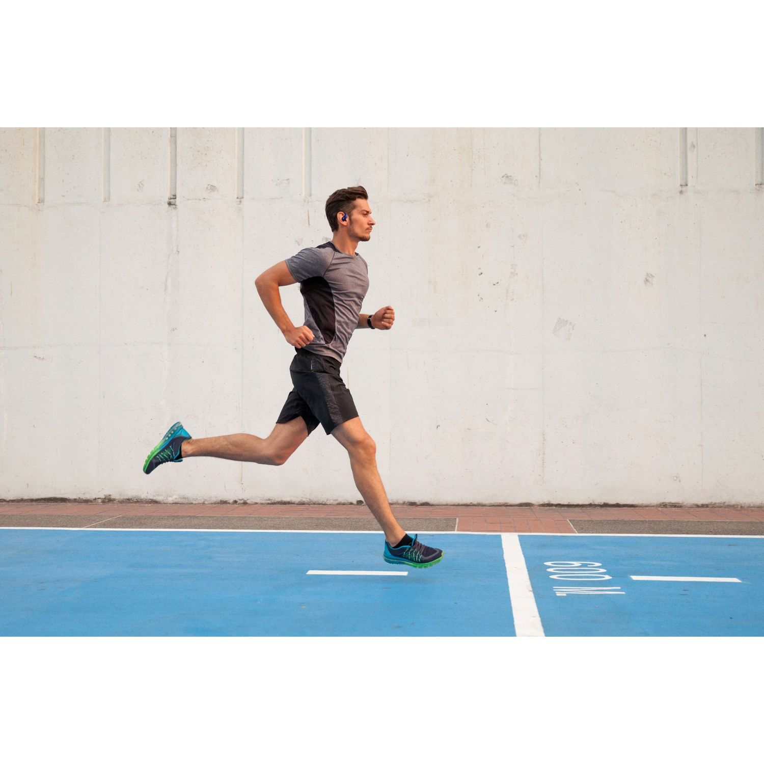 Un homme court sur une piste bleue. Il porte un t-shirt gris, un short noir et des baskets bleues. Un mur blanc est en arrière-plan.
