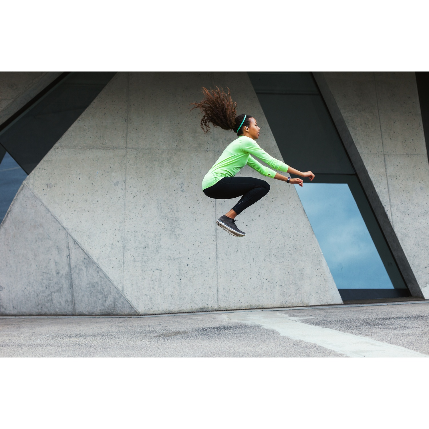 Femme en tenue de sport sautant. Haut vert, leggings noirs et chaussures grises. Bâtiment moderne en arrière-plan.