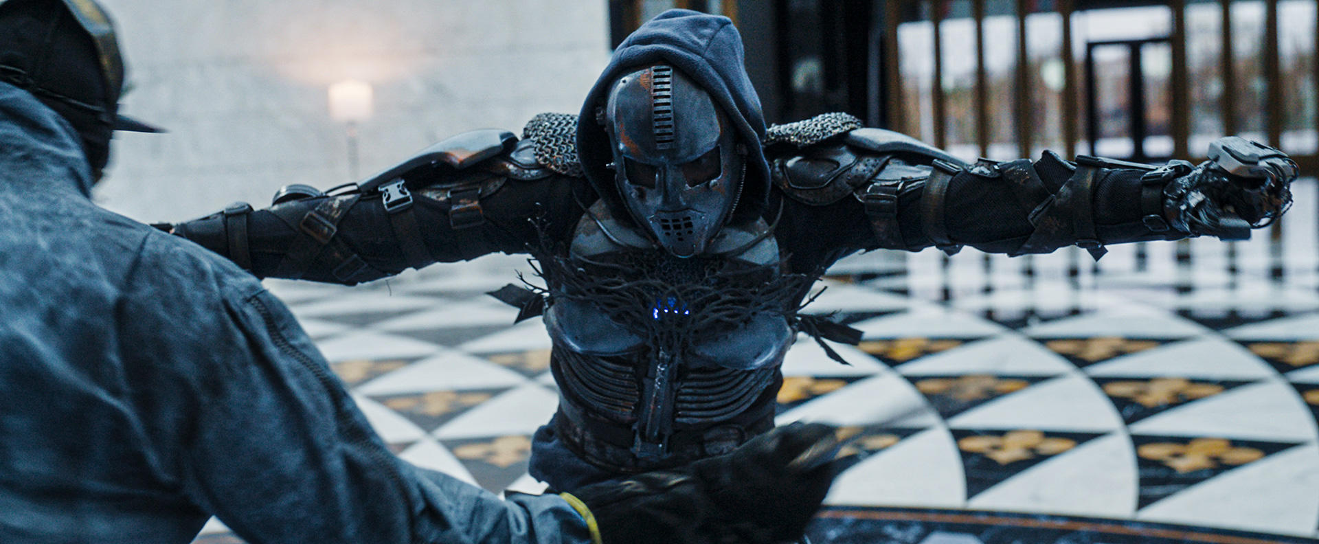 Eine Person in einem futuristischen Anzug mit Helm und Kapuze steht einer anderen Person gegenüber. Beide befinden sich in einem Gebäude mit Schachbrettmuster.
