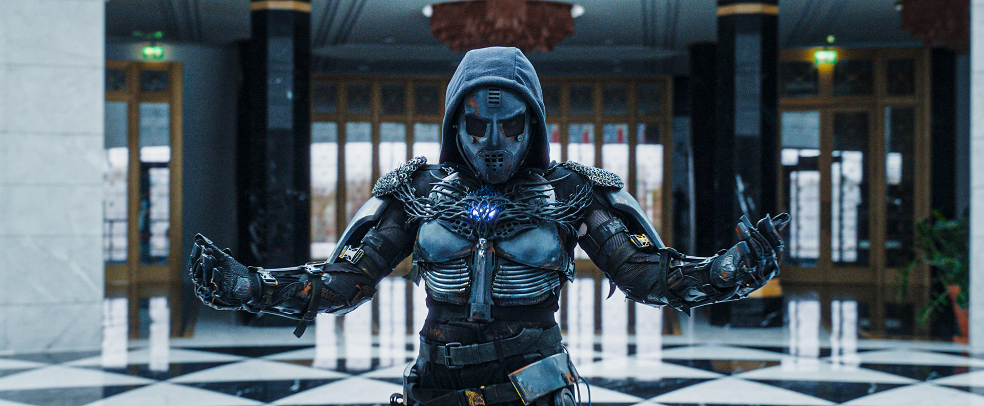 Eine Person in futuristischer Rüstung steht in einem Gebäude. Sie tragen eine Maske und Kapuze. Der Hintergrund zeigt Türen und einen Schachbrettboden.