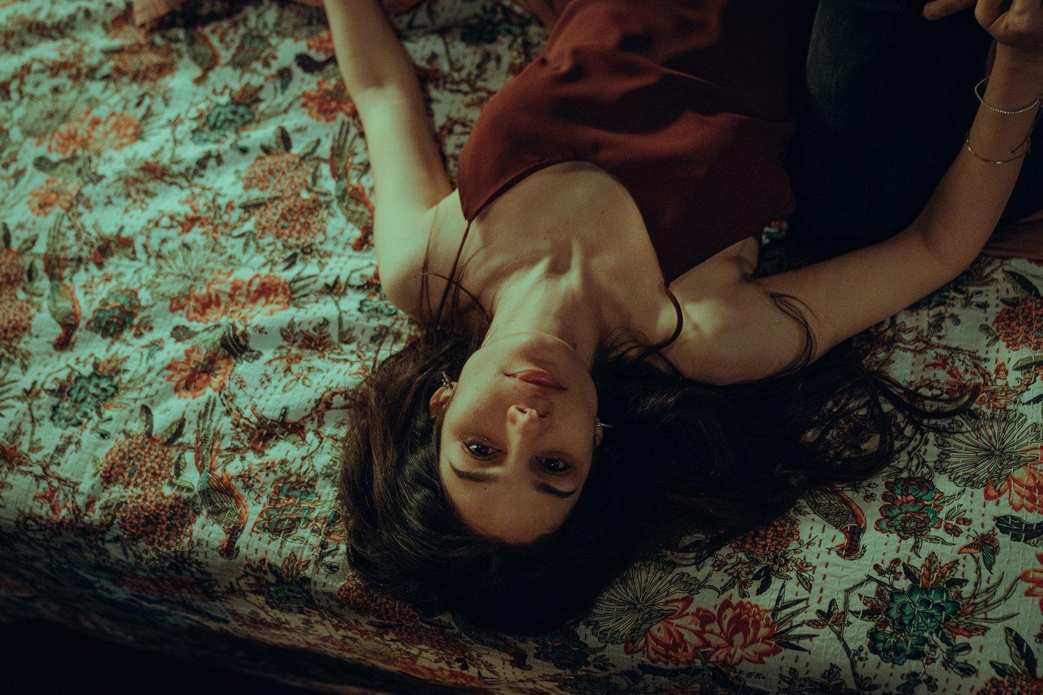 Eine Frau in einem weinroten Kleid liegt auf einem Bett mit einer floral gemusterten Steppdecke und schaut in die Kamera.