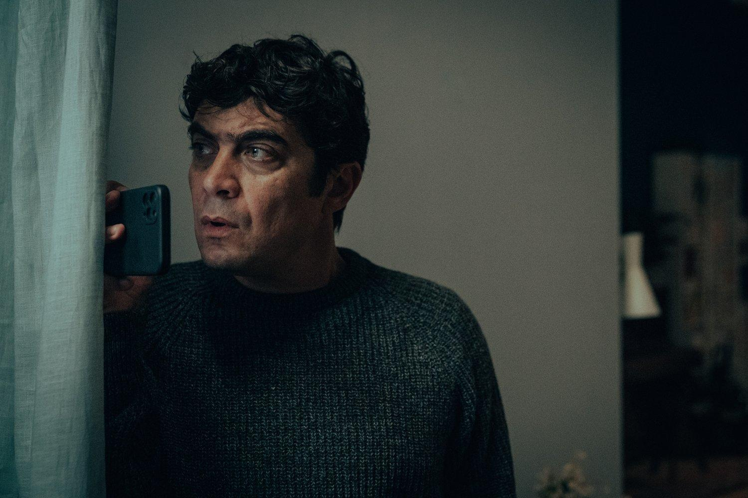 Ein Mann mit Telefon schaut besorgt weg. Er trägt einen dunklen Pullover. Vorhang und Wand sind auf beiden Seiten.