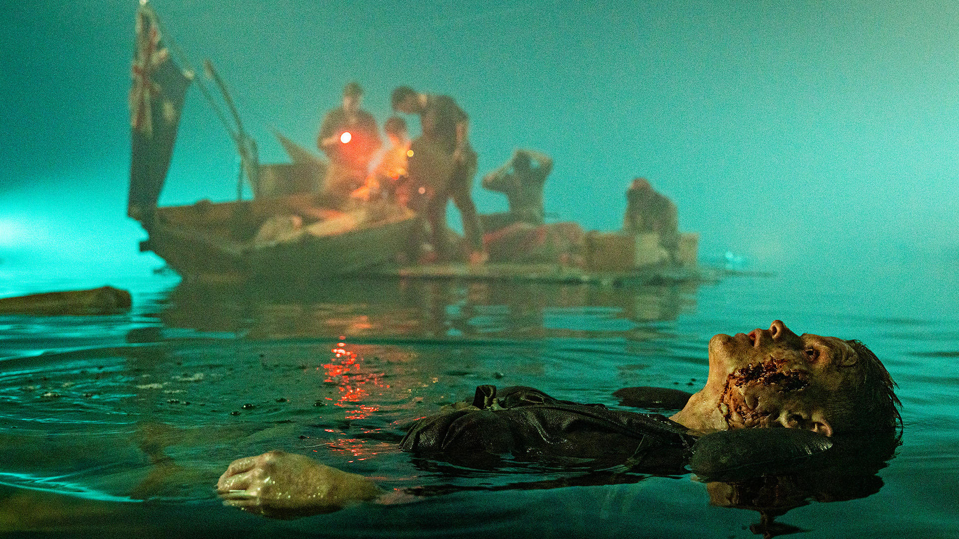 Ein Mann mit Gesichtsverletzungen treibt im Wasser. Ein Boot mit Leuten und Flagge im Hintergrund. Die Szene ist schwach beleuchtet.