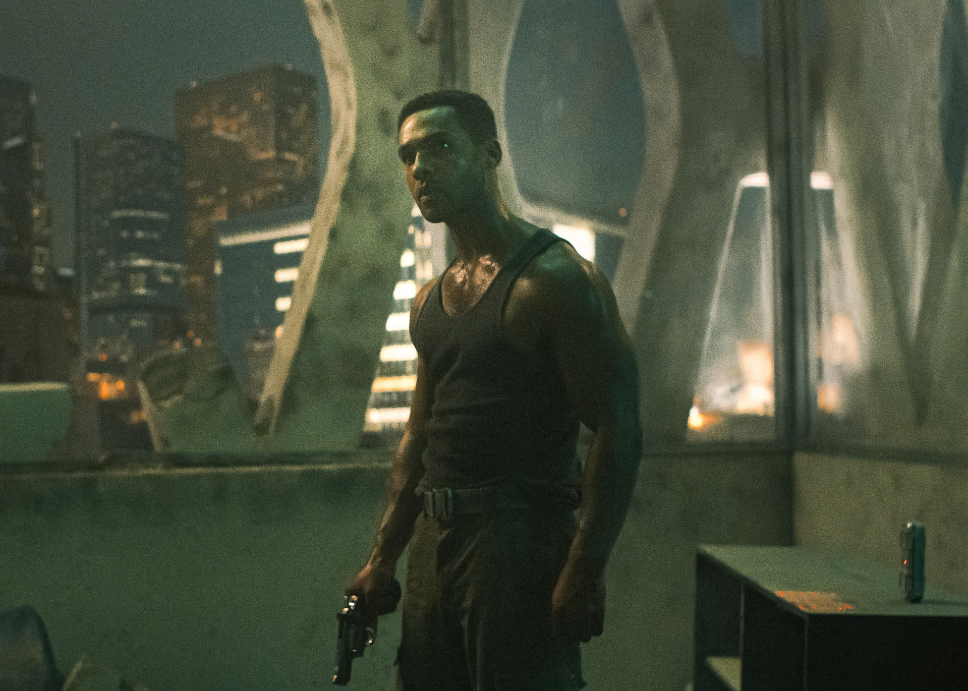 Mann in Tanktop hält eine Pistole und schaut in die Kamera, in einer Stadtlandschaft. Gebäude sind im Hintergrund. Nachtszene.