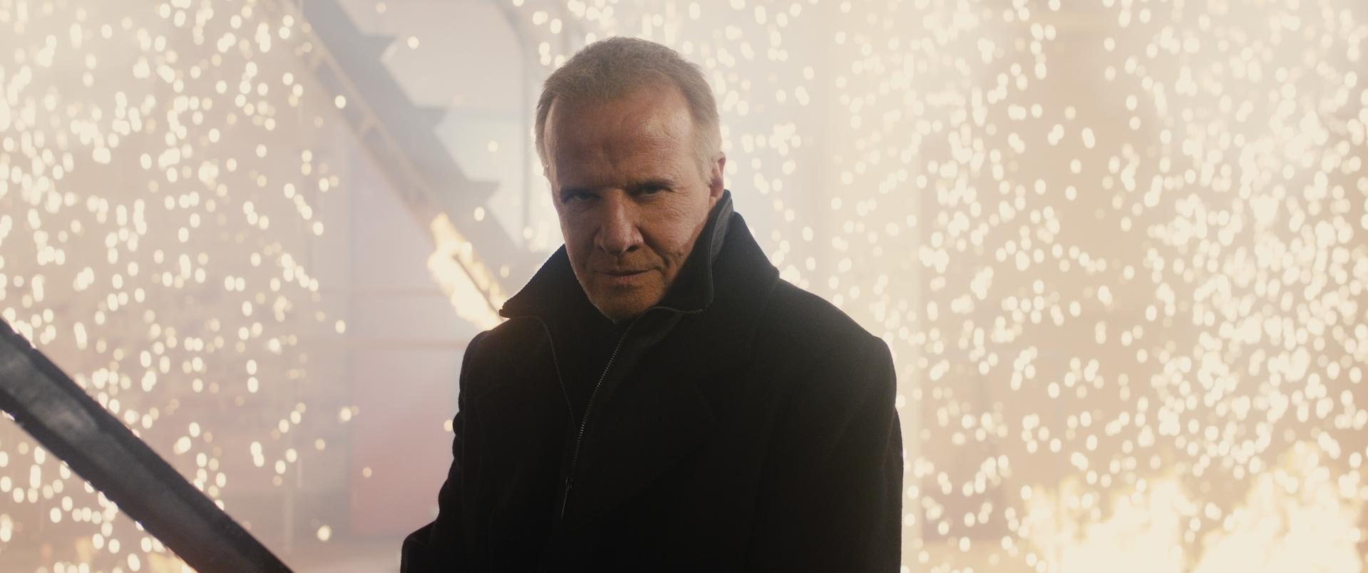 Mann im schwarzen Mantel blickt nach vorne. Der Hintergrund ist mit hellen, verschwommenen Lichtern gefüllt.