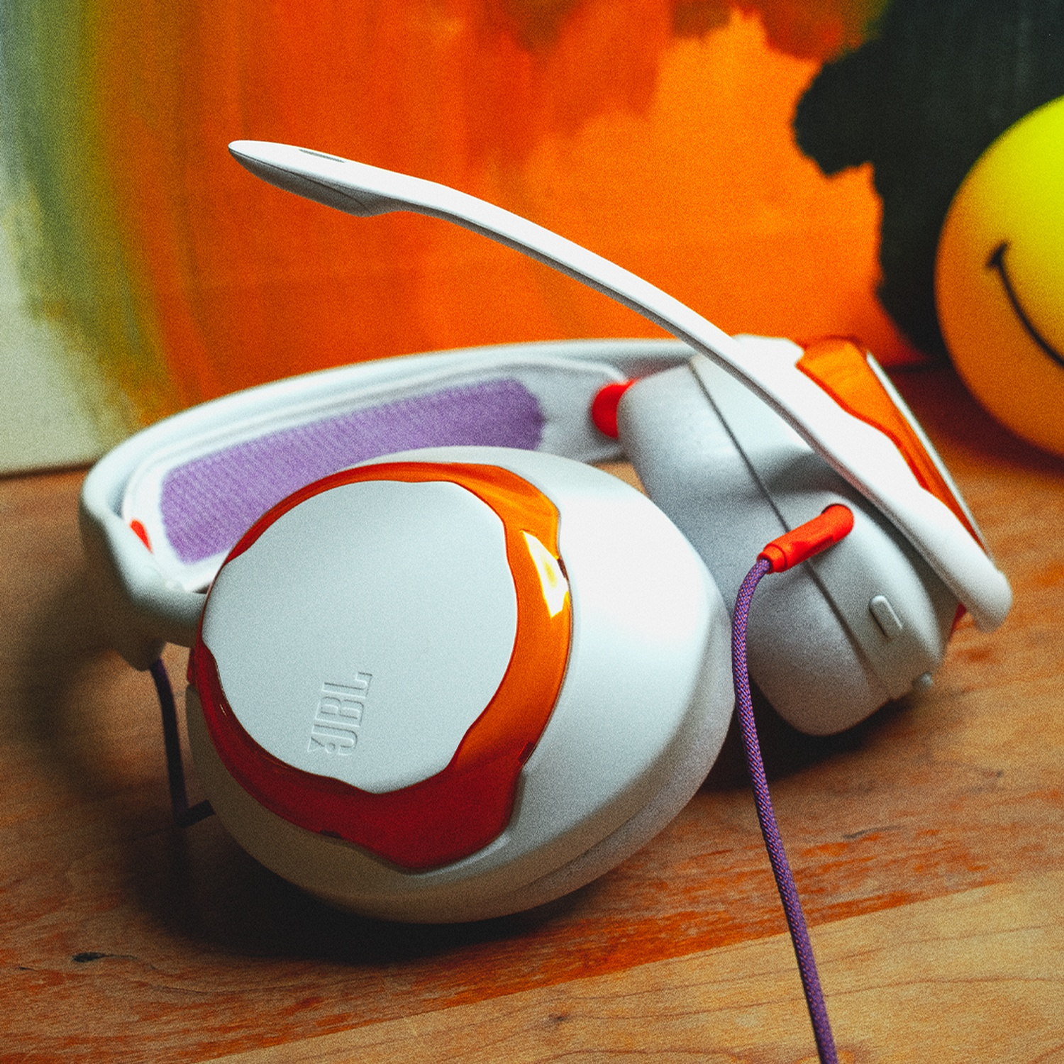 Witte en oranje JBL-koptelefoon met paars snoer op hout. Een oranje en gele achtergrond is zichtbaar.