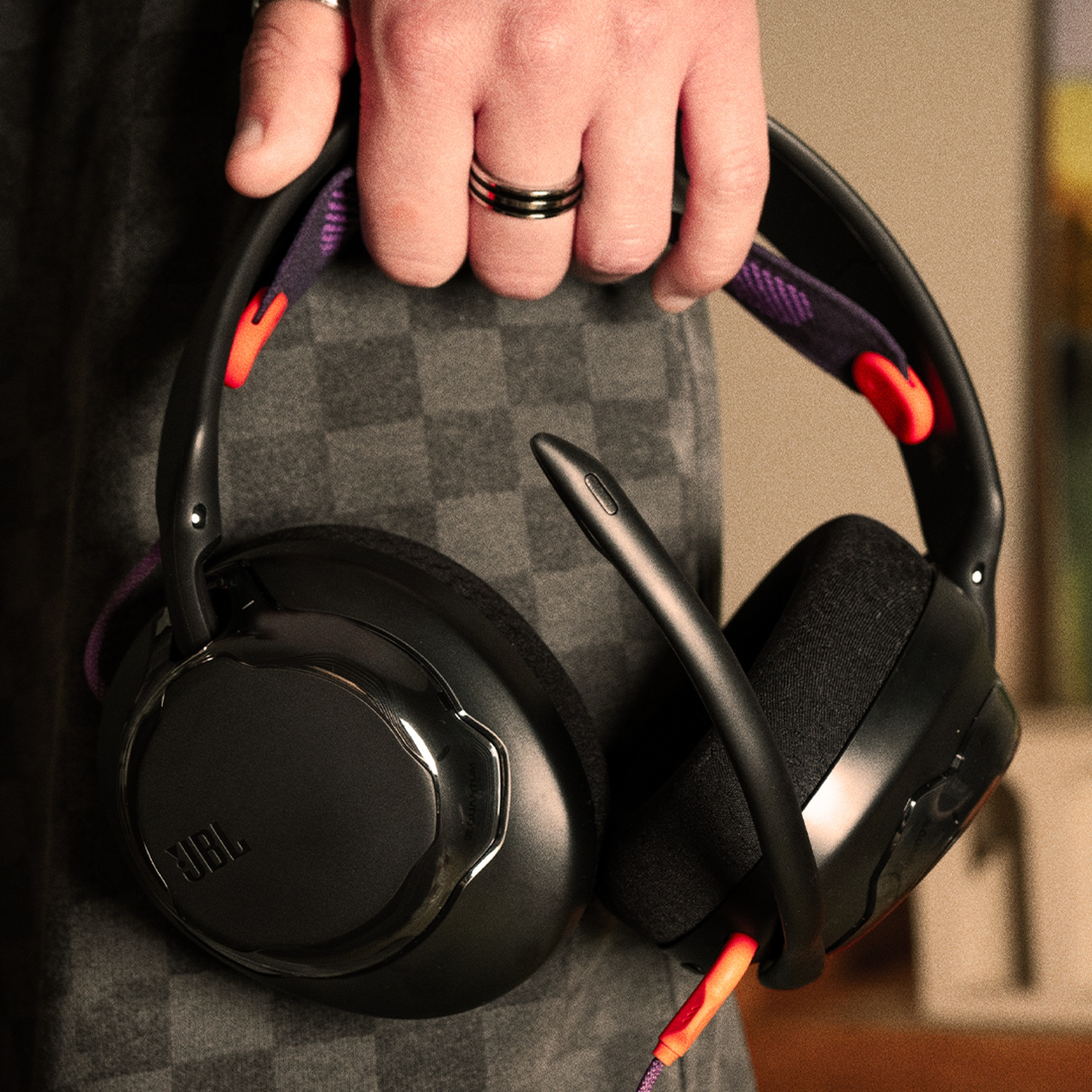 A person holds black JBL headphones with purple and orange accents. The background is a gray checkered fabric and a blurred room.