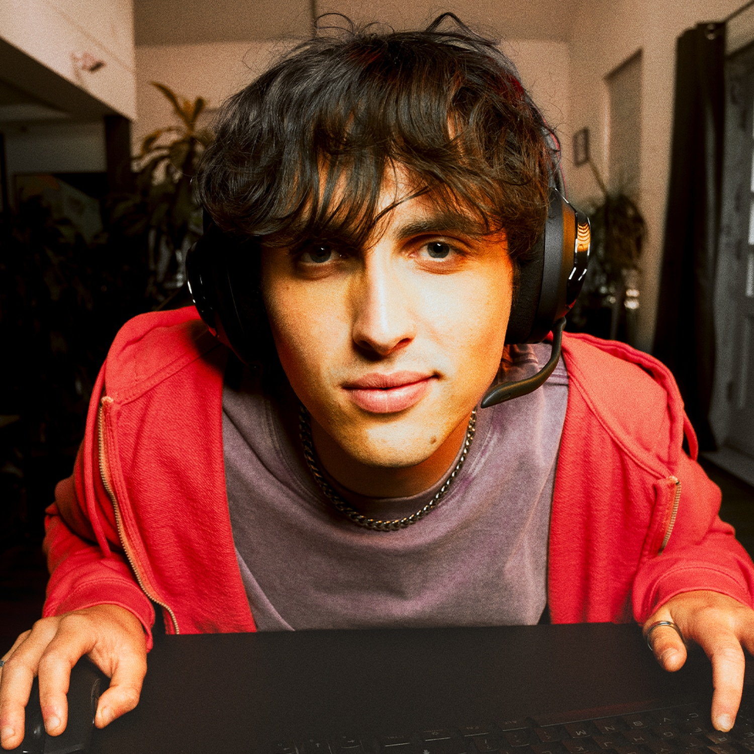 Un jeune homme portant un casque et une veste rouge regarde la caméra. Il est à un bureau avec un clavier. Arrière-plan : plantes et une fenêtre.