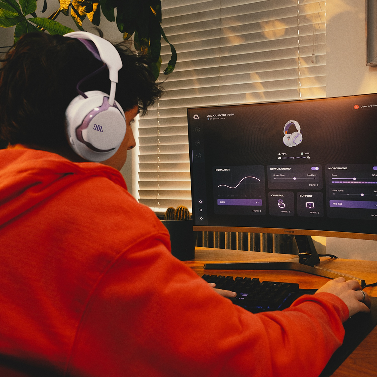 Personne portant un casque JBL, utilisant un ordinateur. L'écran affiche les paramètres audio. Un sweat à capuche orange est porté, avec un cactus en arrière-plan.
