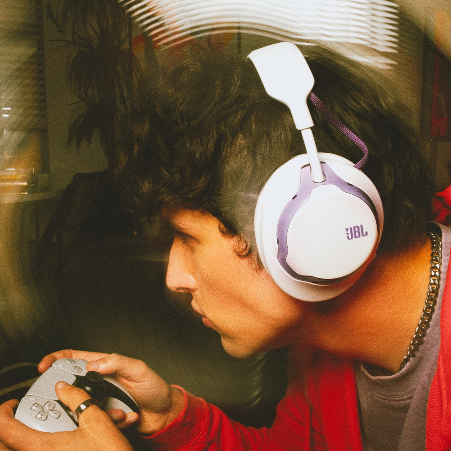 Une personne portant un casque JBL blanc joue avec une manette. Elle porte une veste rouge et une chaîne argentée.