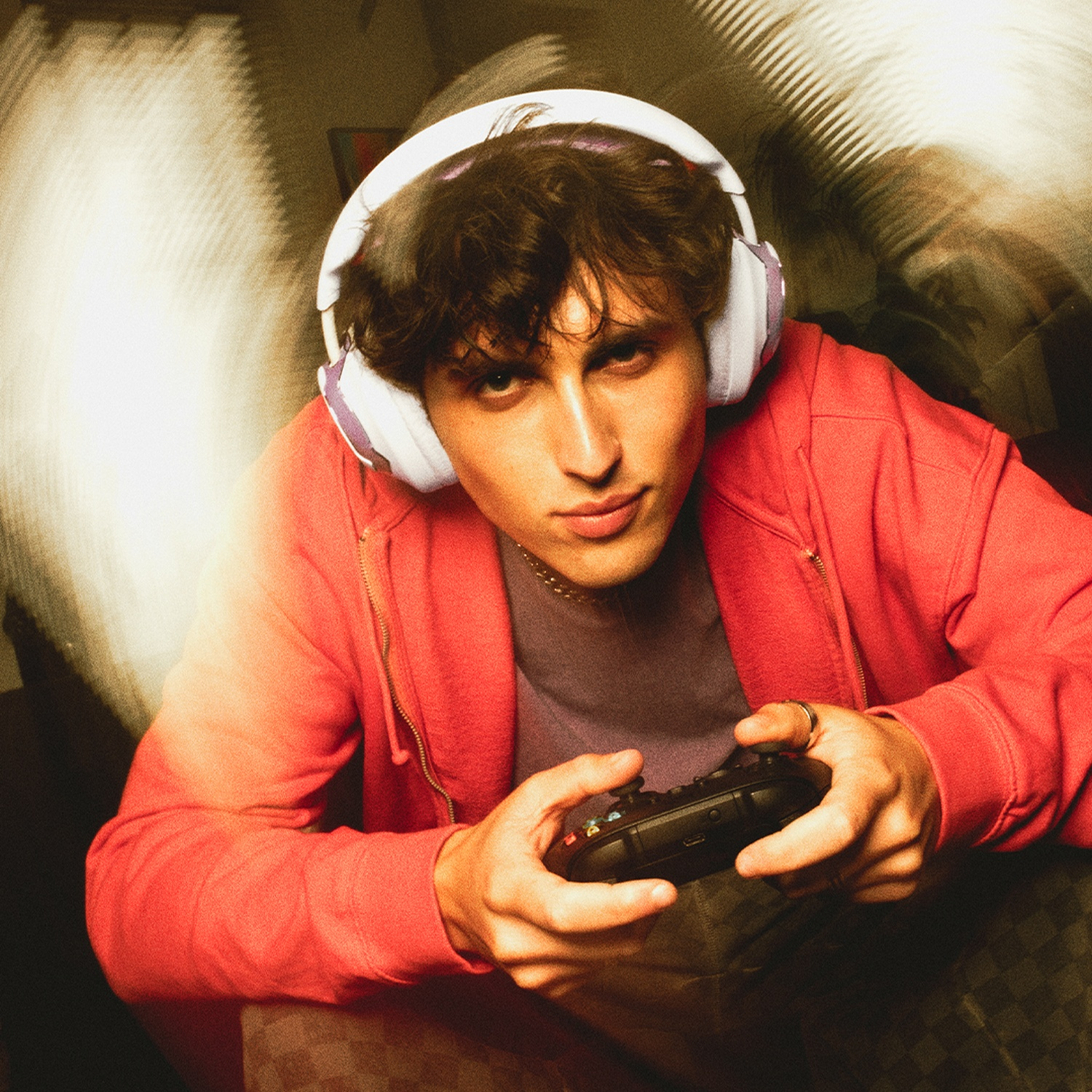 Un jeune homme portant un casque et un sweat à capuche rouge tient une manette de jeu. Il regarde la caméra, assis dans une pièce.