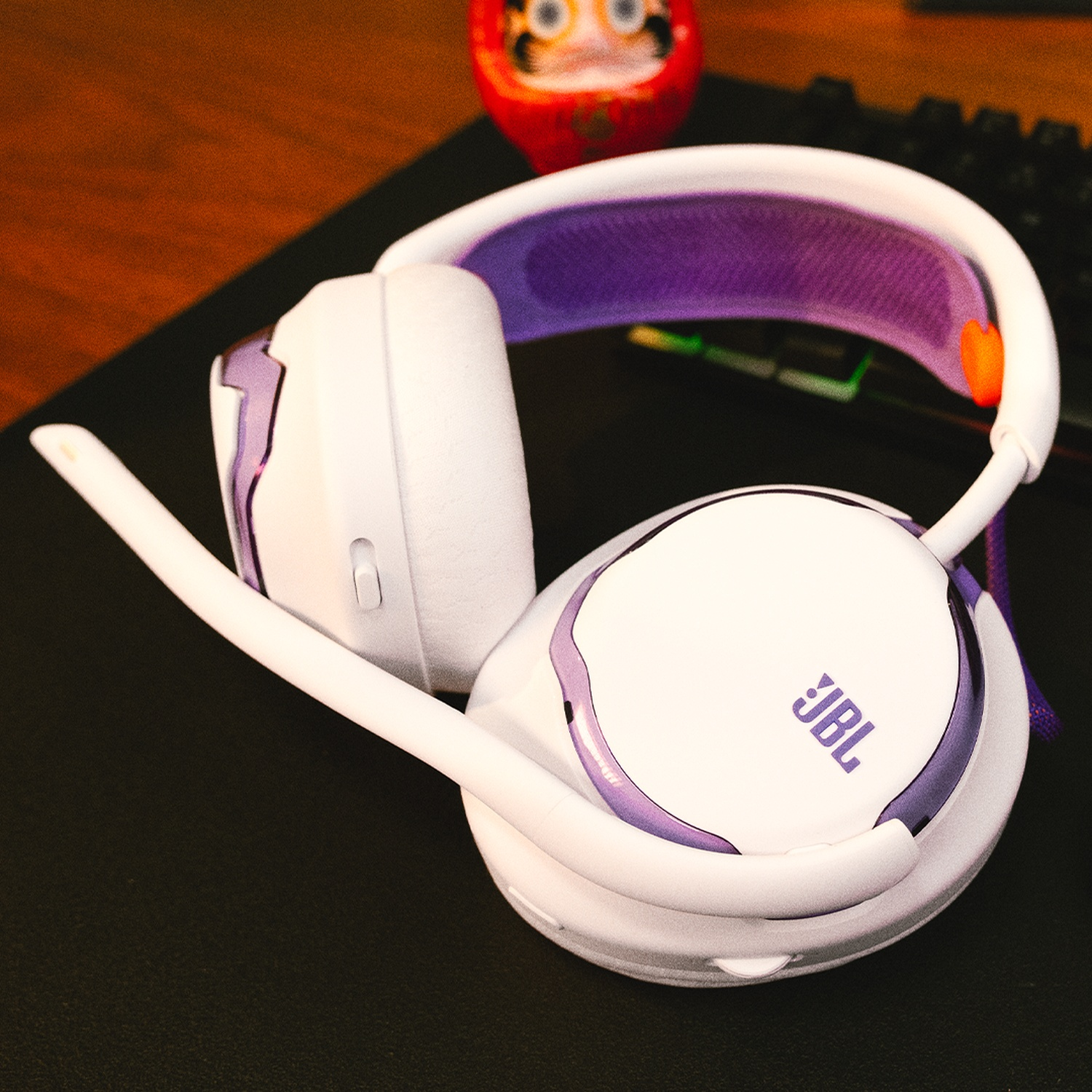 Un casque JBL blanc avec des accents violets repose sur une surface noire. Une poupée Daruma rouge est en arrière-plan.