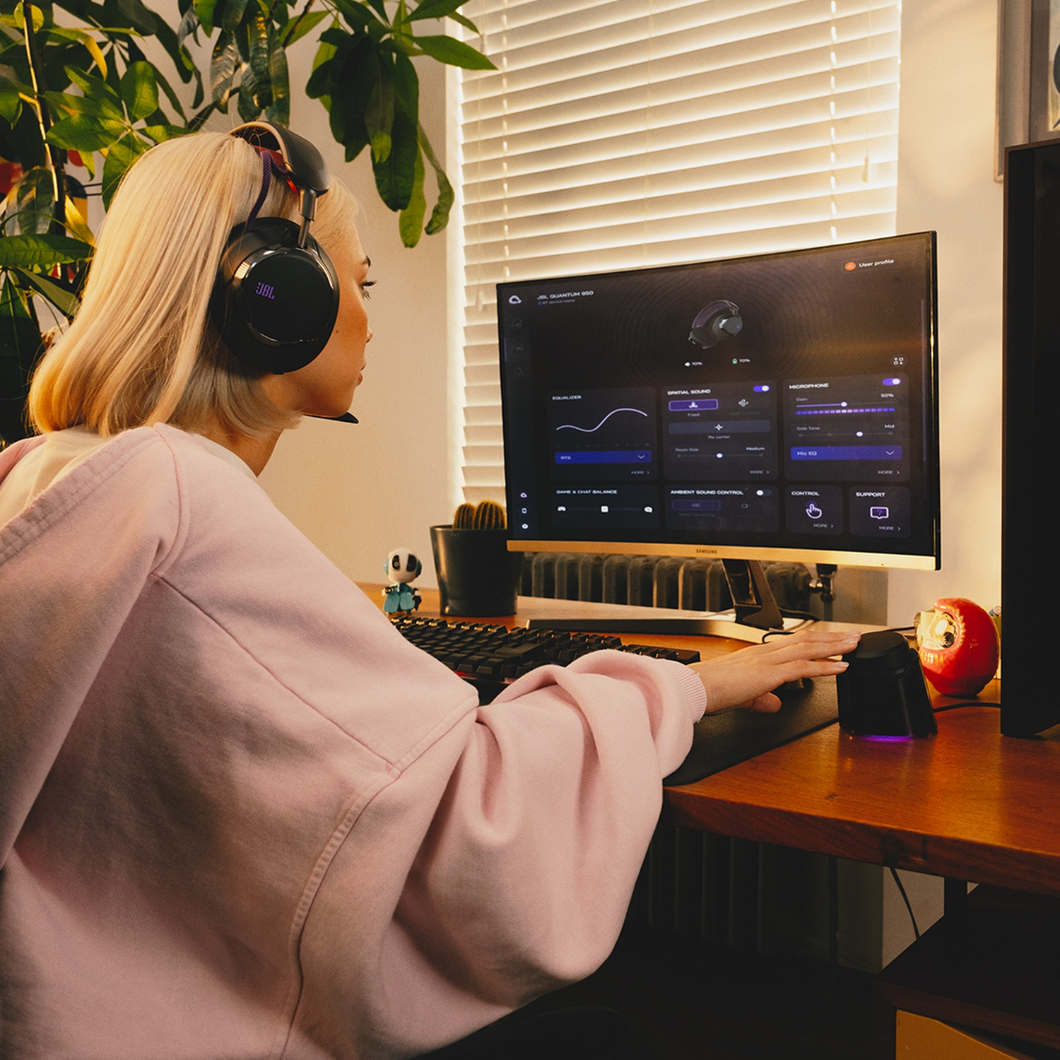 Een vrouw met een koptelefoon zit aan een bureau en gebruikt een computer. Een monitor toont een geluidsinstellingeninterface. Een luidspreker staat op het bureau.