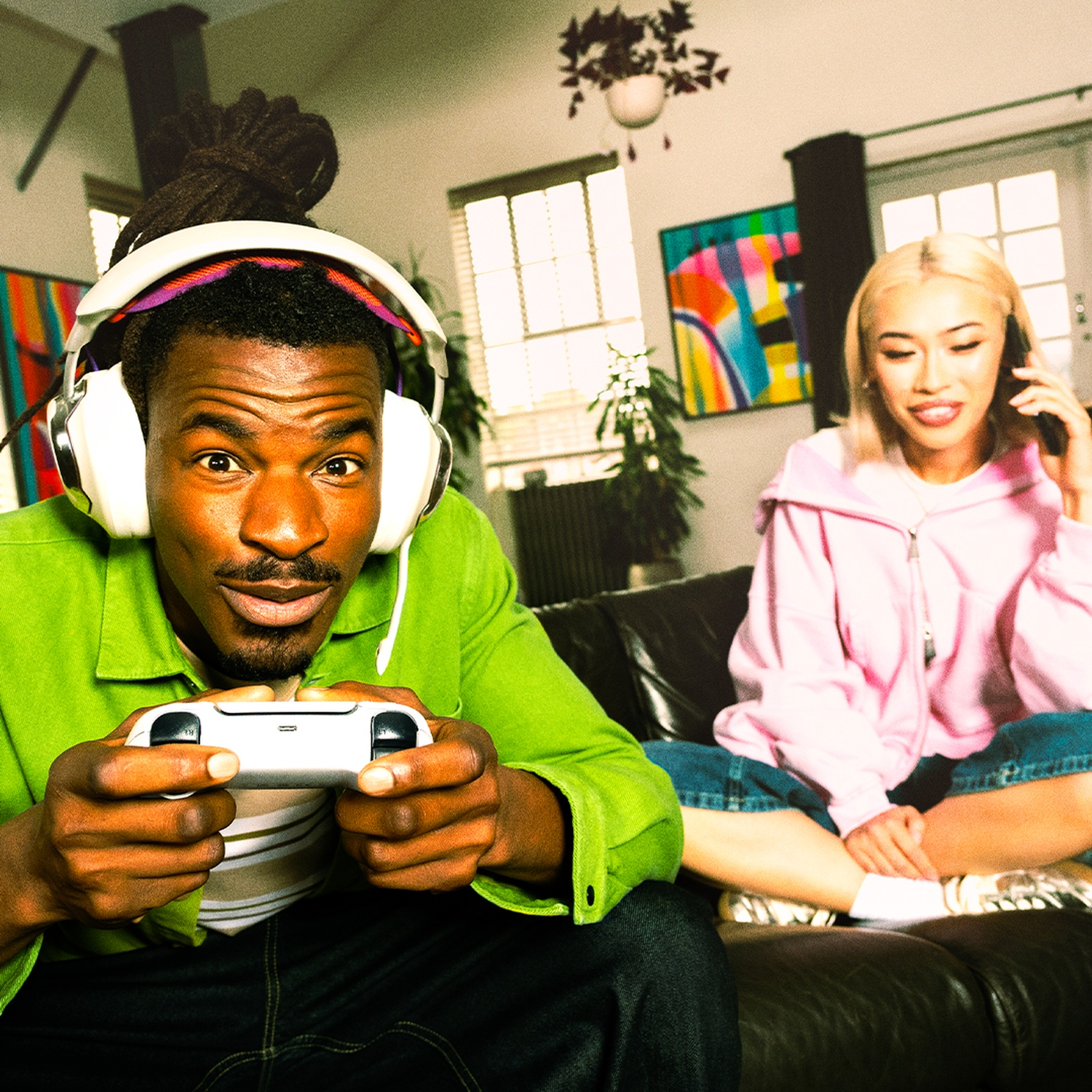 Een man met een koptelefoon speelt een videogame. Een vrouw zit op een bank en belt. De scène speelt zich binnenshuis af.