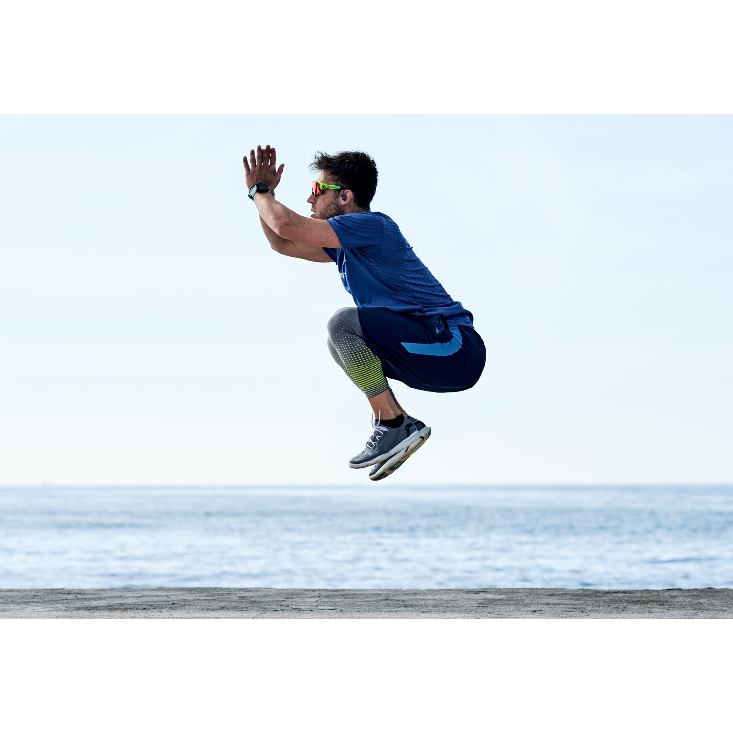 Un homme saute en l'air près de la mer. Il porte des lunettes de soleil, une chemise bleue et des chaussures grises. L'arrière-plan montre la mer et le ciel.
