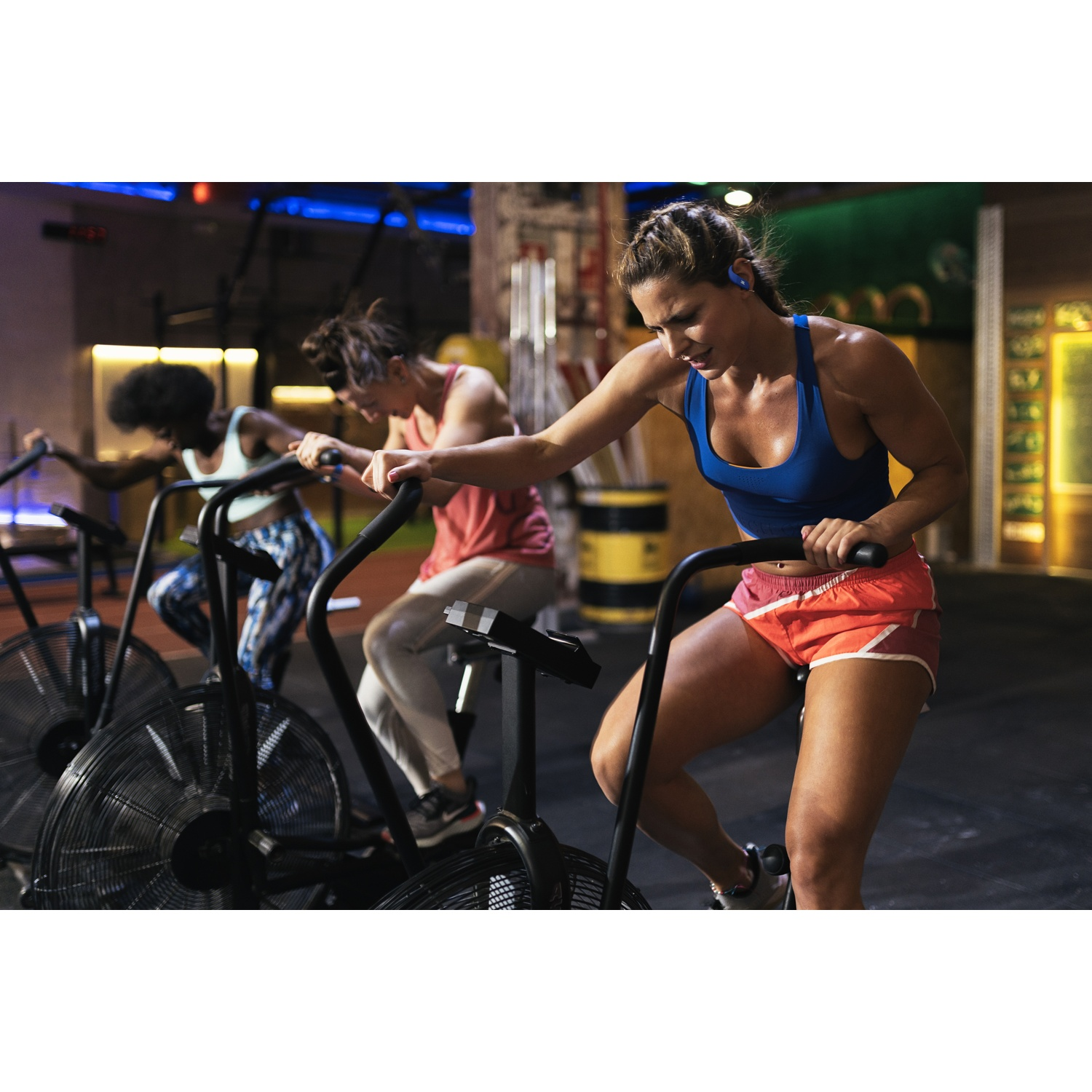 Trois femmes sur des vélos d'exercice dans une salle de sport. L'accent est mis sur une femme en haut bleu et short orange.