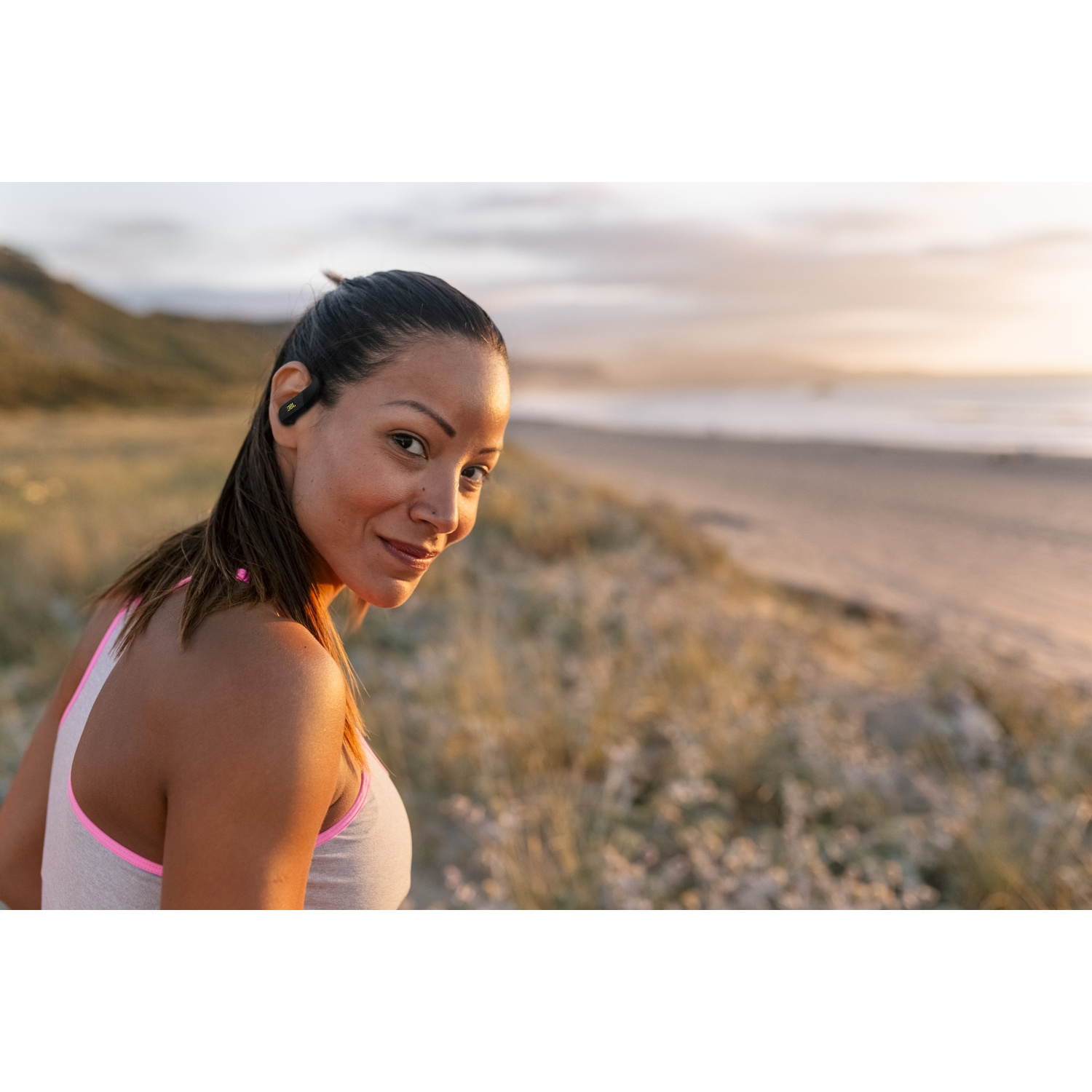 Femme portant des écouteurs sur une plage, regardant la caméra. Elle porte un débardeur gris avec une bordure rose.