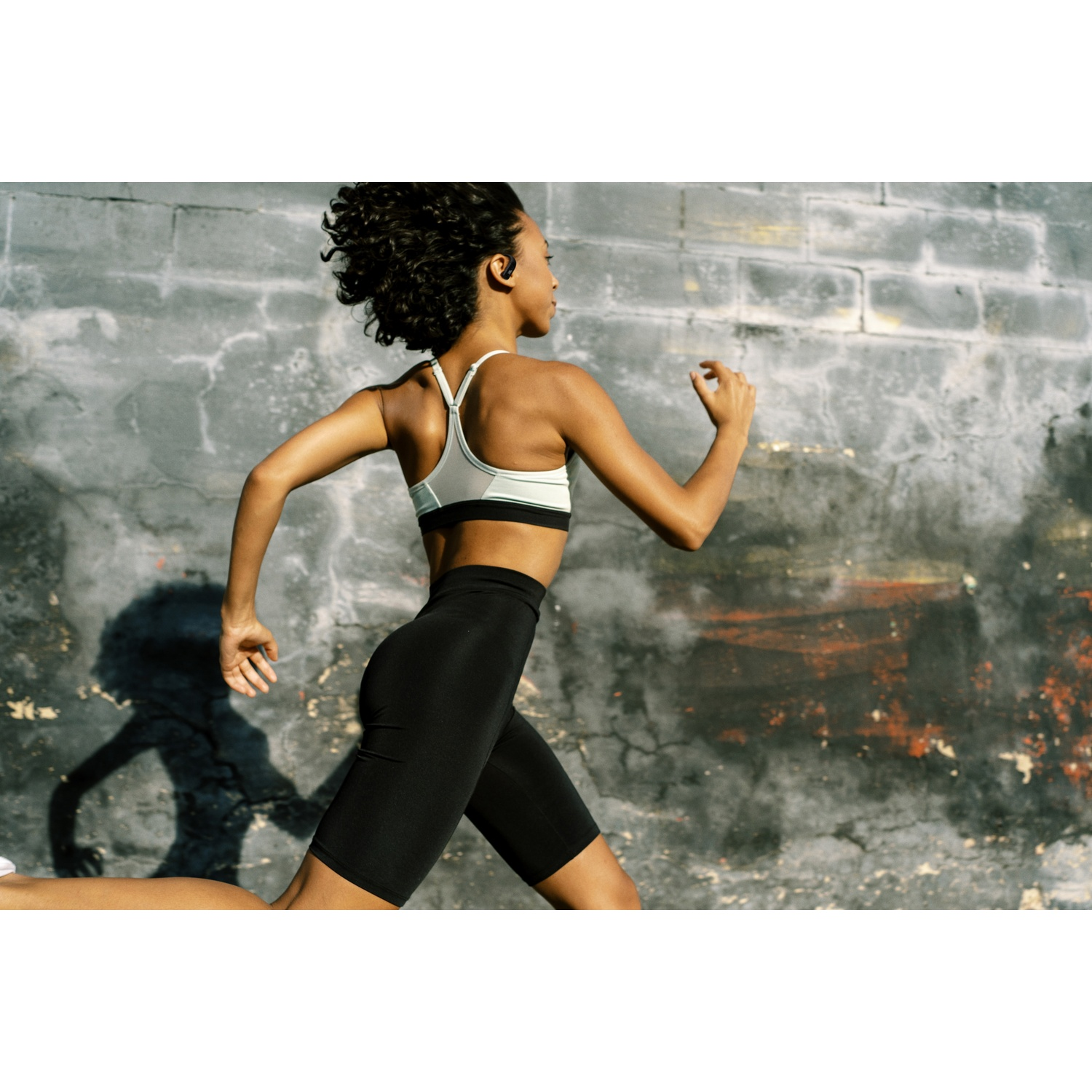 Une femme court devant un mur usé. Elle porte un soutien-gorge de sport et un short, avec des écouteurs. Son ombre est visible sur le mur.