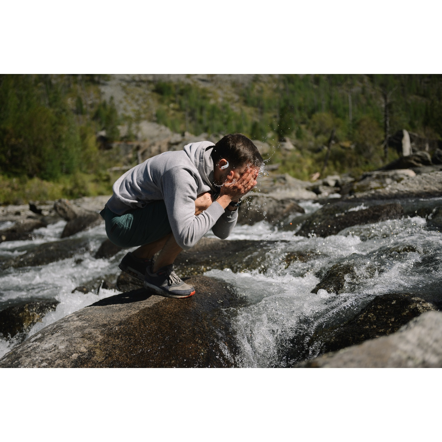 Un homme se lave le visage avec l'eau d'une rivière. Il porte un sweat à capuche gris et un short. L'arrière-plan est une forêt.