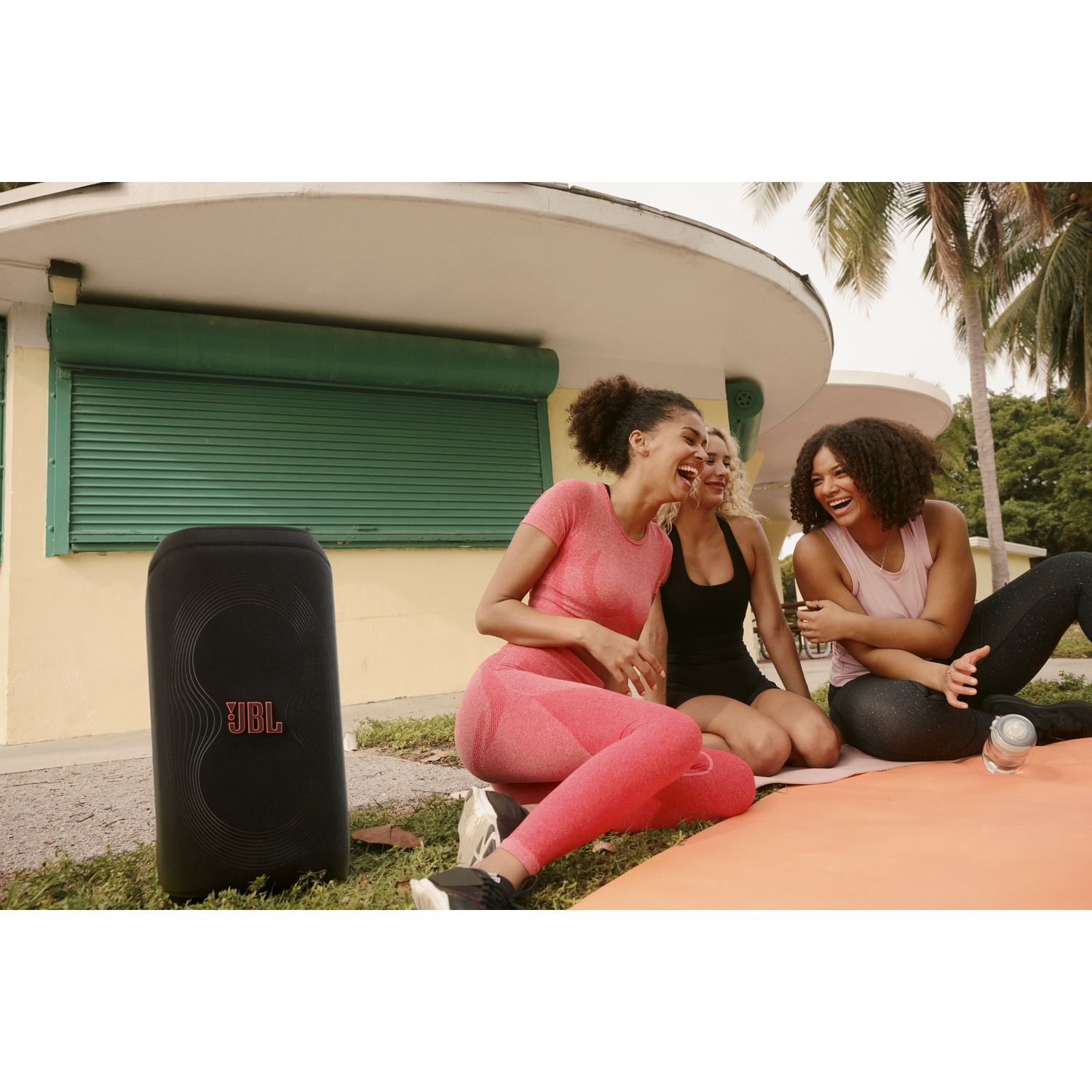 Drie vrouwen lachen, zittend op een oranje mat bij een zwarte JBL-luidspreker. Een draagt roze, een zwart, en een lichtroze top.