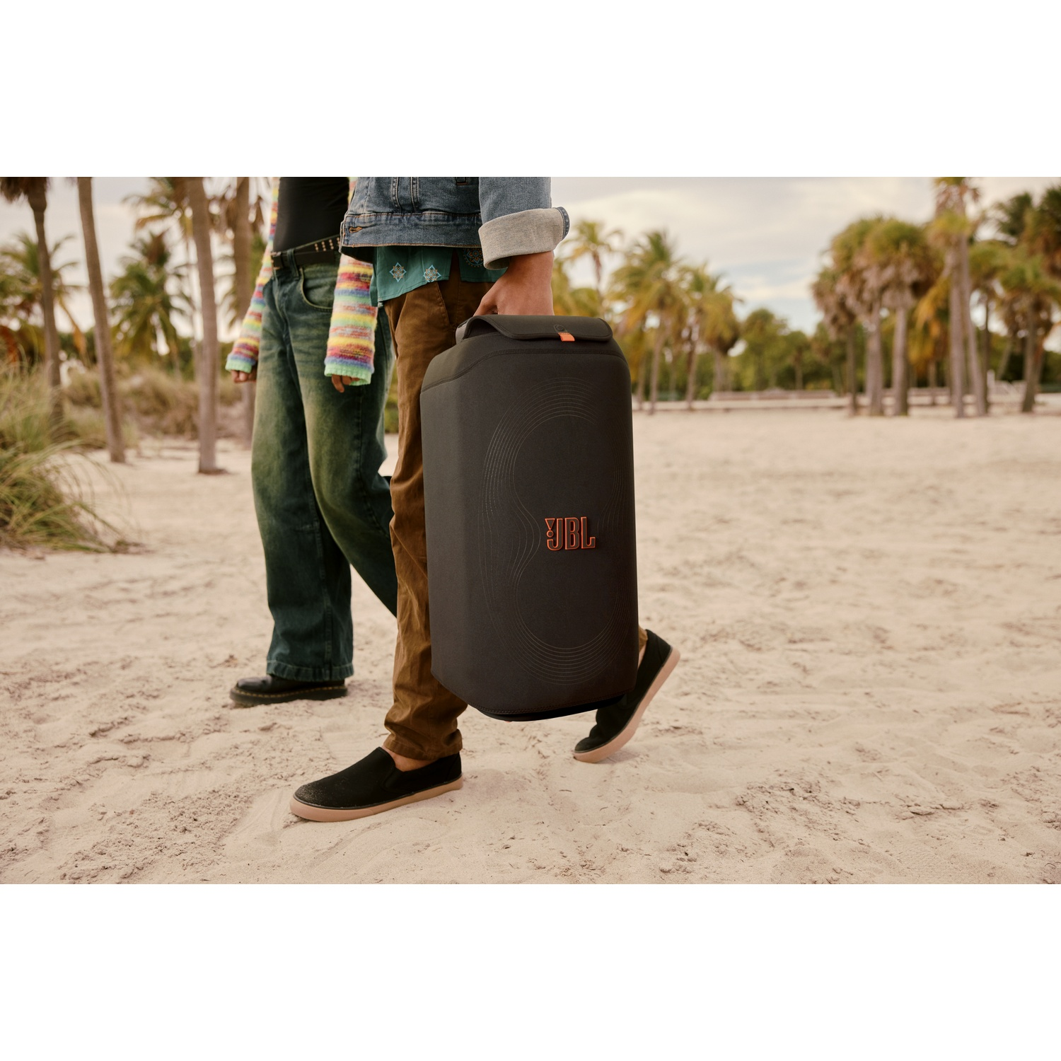 Twee mensen lopen op een strand, een draagt een zwarte JBL-luidspreker. Palmbomen op de achtergrond.