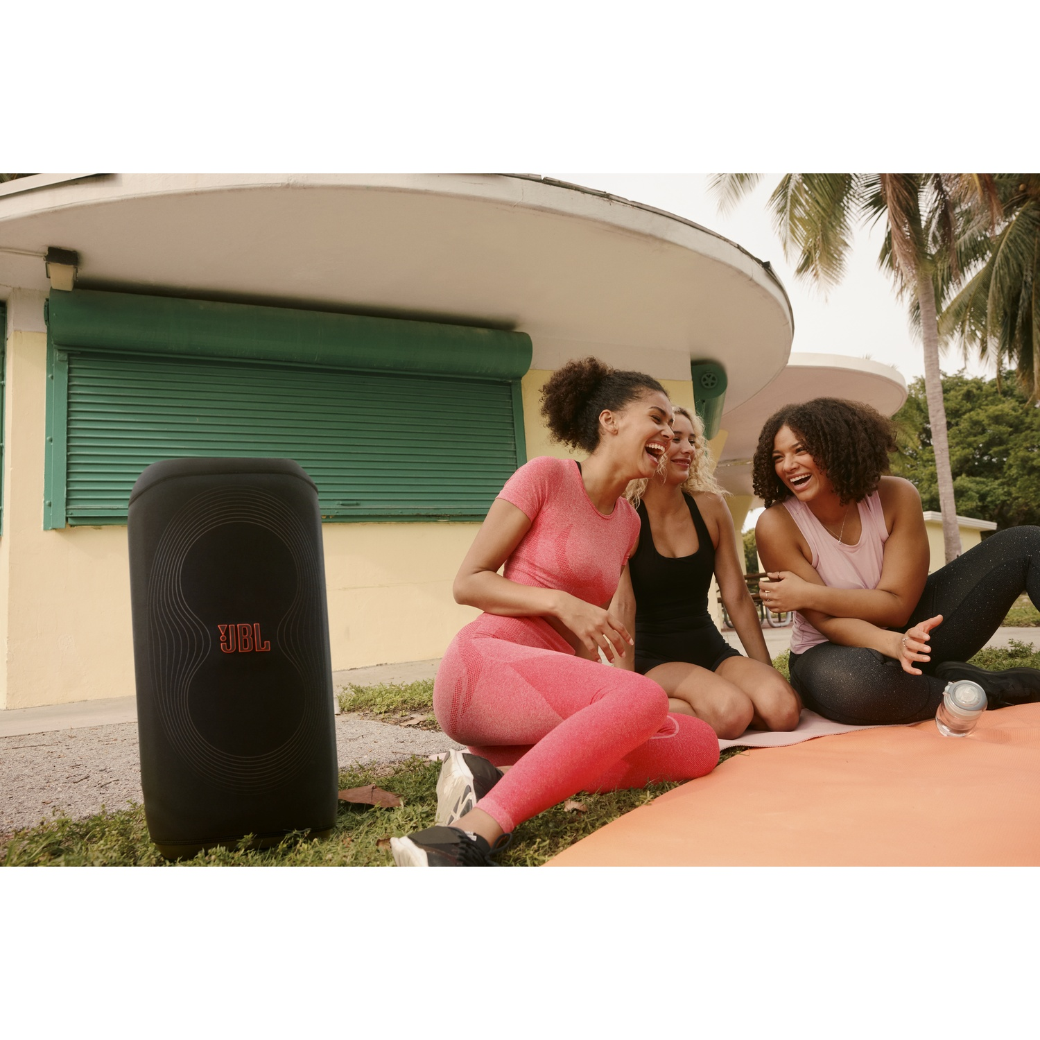 Drie vrouwen lachen terwijl ze op een mat zitten. Een zwarte JBL-luidspreker staat op gras. Een gebouw en palmbomen op de achtergrond.