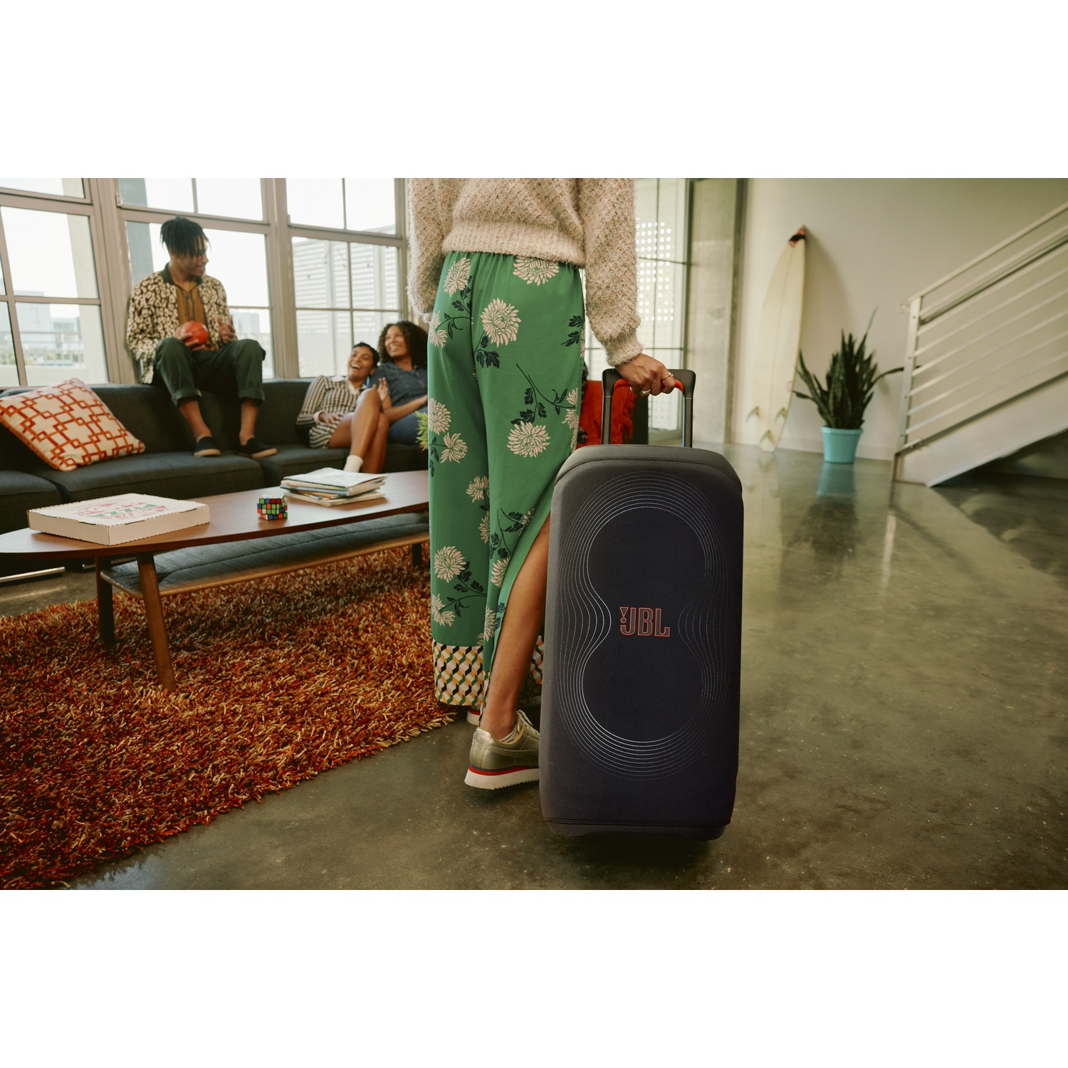 Een persoon houdt een JBL-luidspreker vast in een kamer. Anderen zitten op een bank. Een surfplank en een plant staan op de achtergrond.