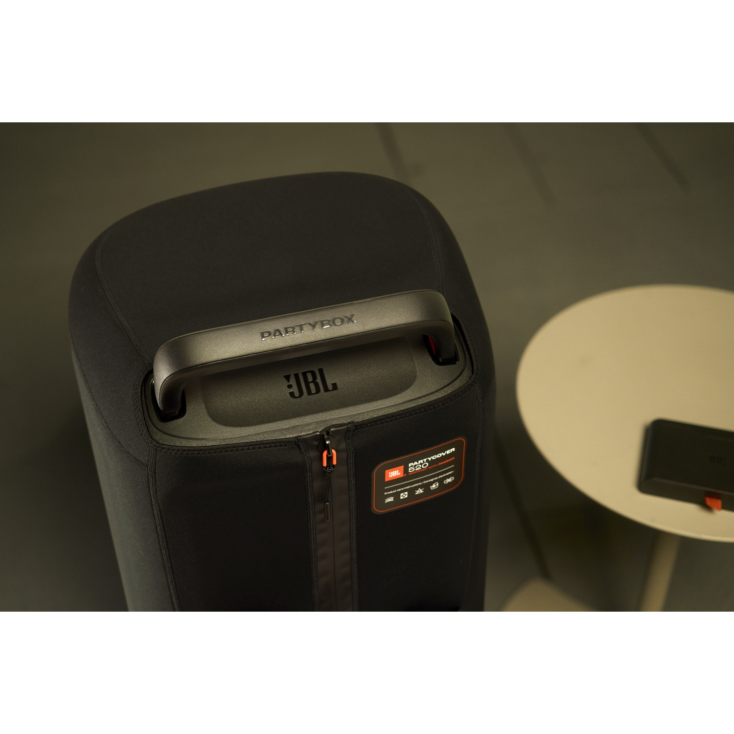 Een zwarte JBL PartyBox-luidspreker met een handvat. Een kleine tafel en een apparaat staan op de achtergrond. De scène is binnen.