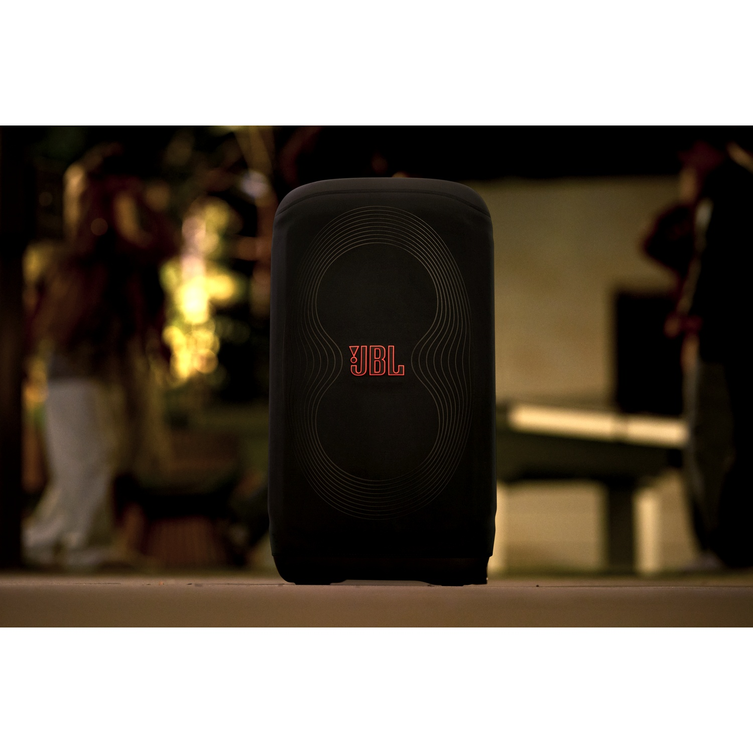 Een zwarte JBL-luidspreker staat in het midden, met rood JBL-logo. Mensen zijn wazig op de achtergrond, met warme omgevingsverlichting.