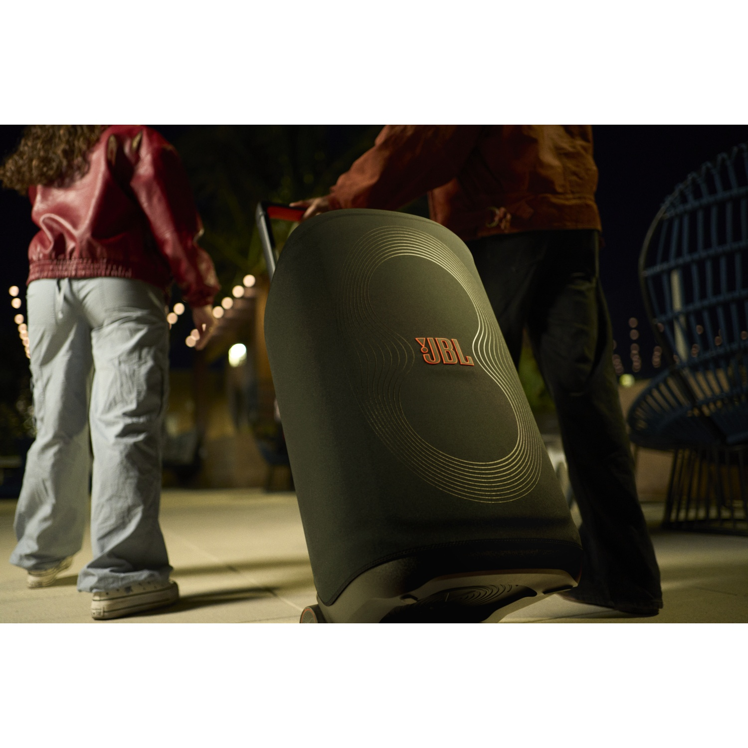 Mensen trekken aan een zwarte JBL-luidspreker. Eén persoon draagt een rode jas. De luidspreker heeft het JBL-logo. Nachtscène met lichtslingers.