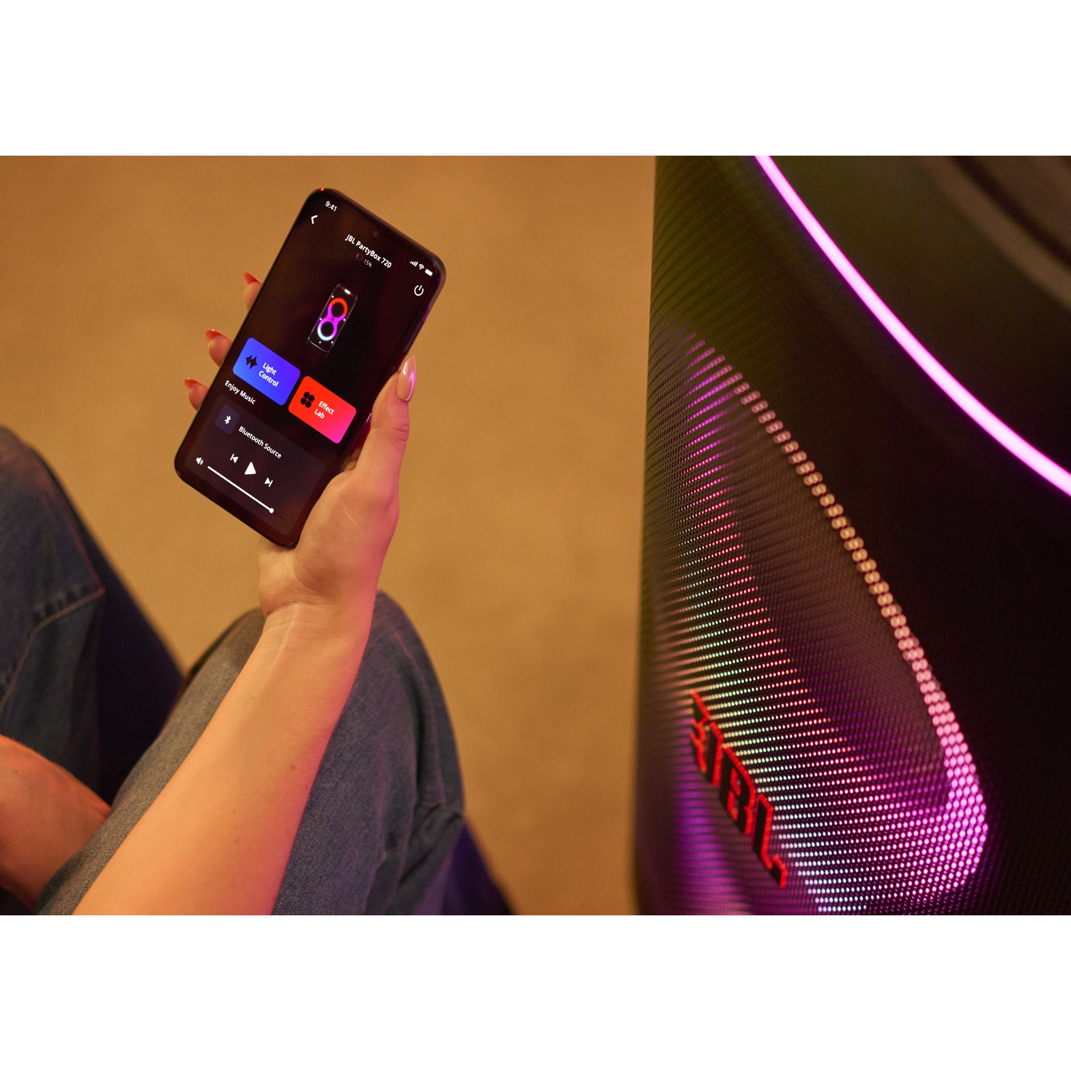Een hand houdt een telefoon met een muziek-app vast, in de buurt van een luidspreker met kleurrijke lichten. Het scherm toont bedieningselementen.