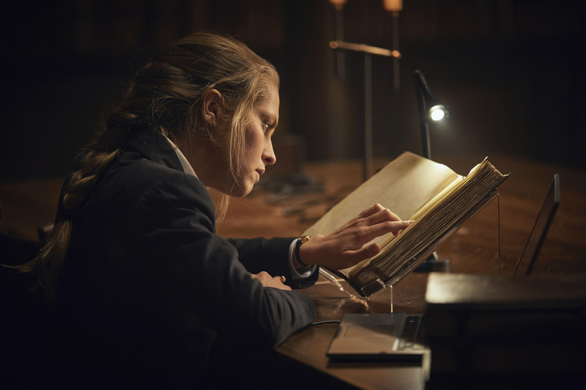 Eine Frau liest ein großes Buch unter einer Lampe. Sie trägt eine dunkle Jacke, der Raum ist schwach beleuchtet.
