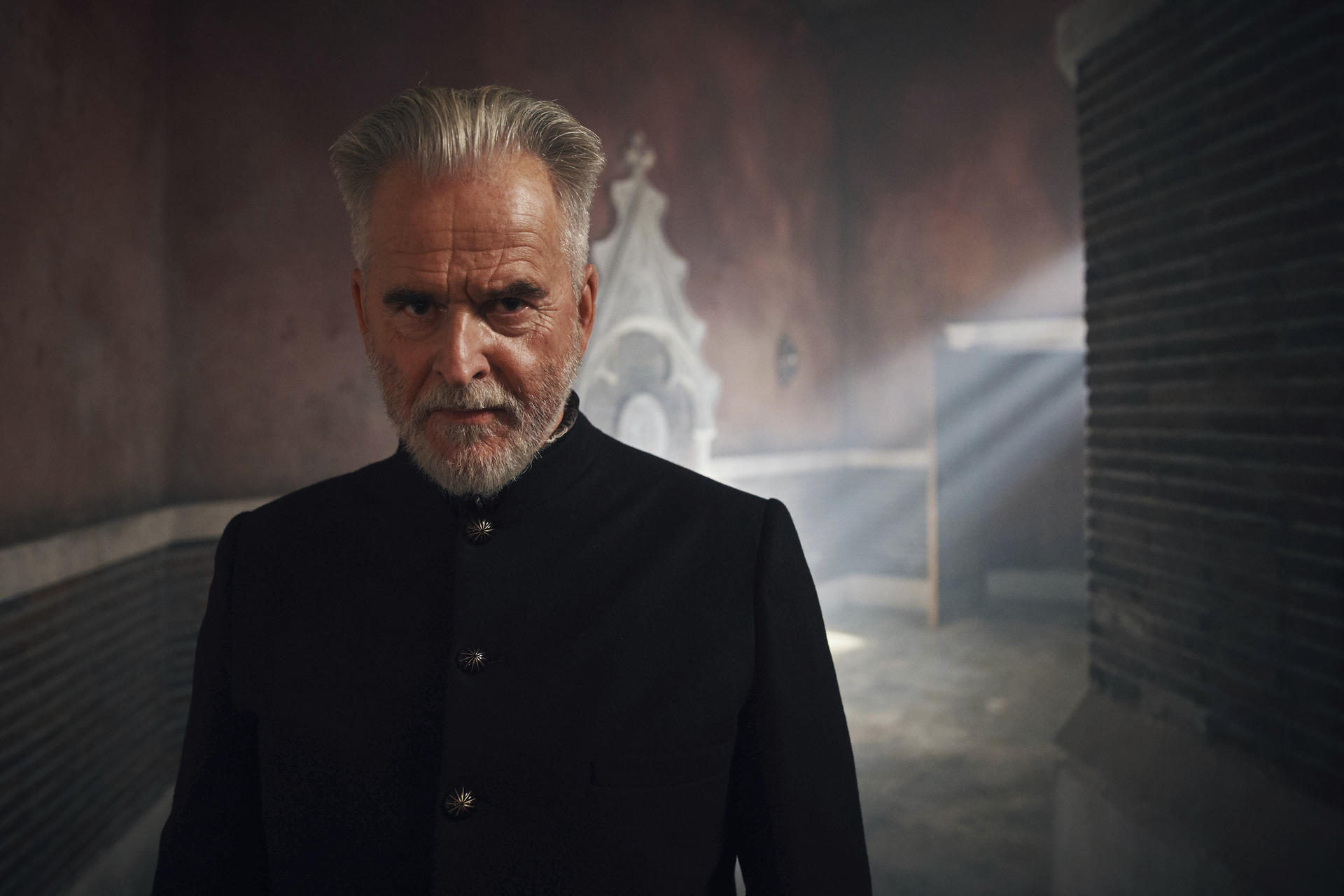 Ein älterer Mann mit grauem Bart und Haar, trägt eine schwarze Jacke und starrt intensiv. Schwach beleuchteter Raum, Lichtstrahlen im Hintergrund sichtbar.