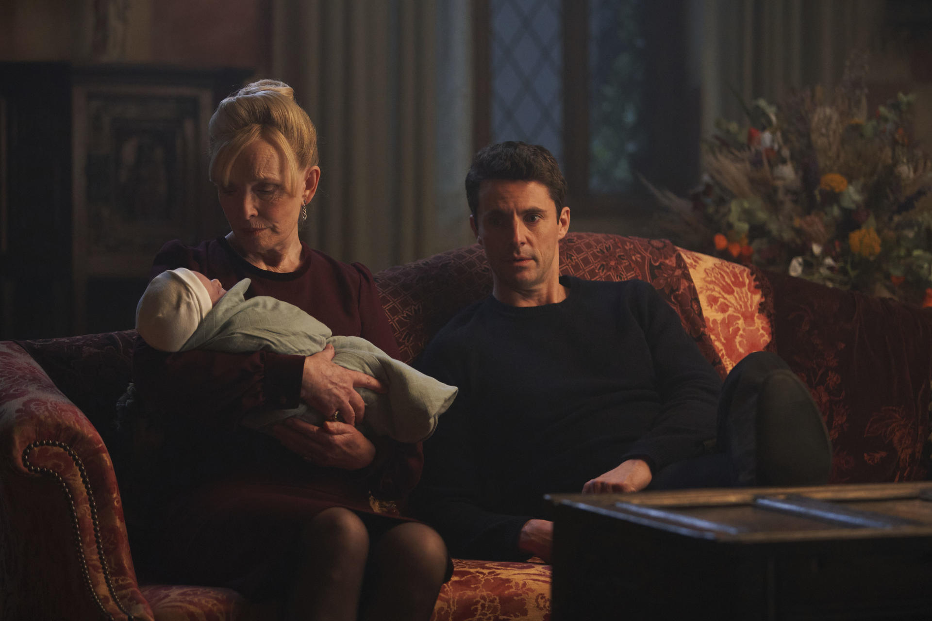 Eine Frau hält ein Baby. Ein Mann sitzt neben ihr. Sie sitzen auf einer floralen Couch in einem schwach beleuchteten Raum. Blumen sind im Hintergrund.