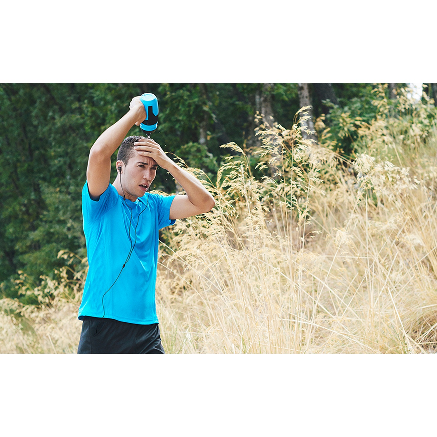 Un homme en chemise bleue se verse de l'eau sur la tête avec une bouteille. Il est en extérieur, avec des herbes hautes et des arbres en arrière-plan.