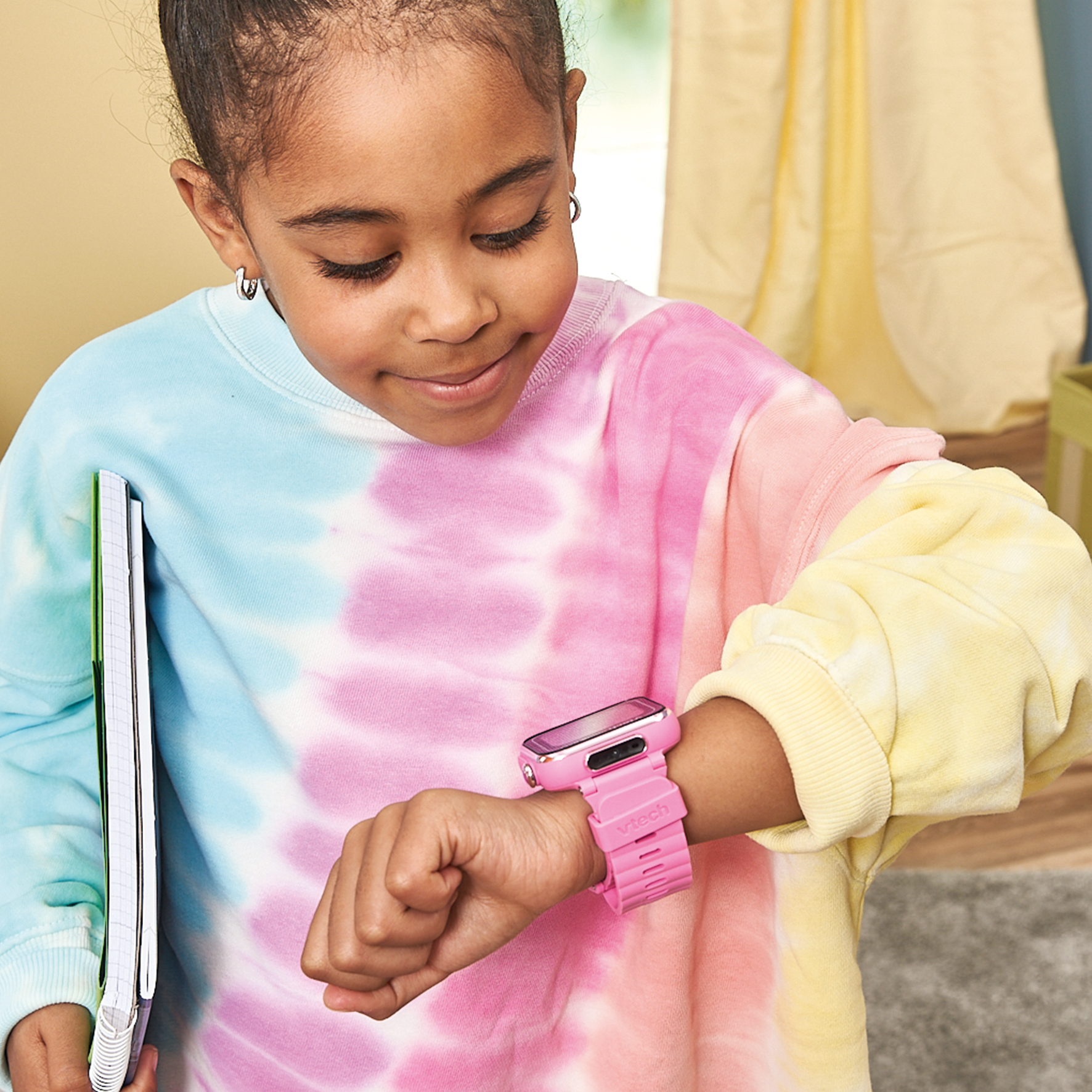 Una niña mira un reloj rosa en su muñeca. Sostiene un cuaderno. Lleva un suéter teñido anudado.