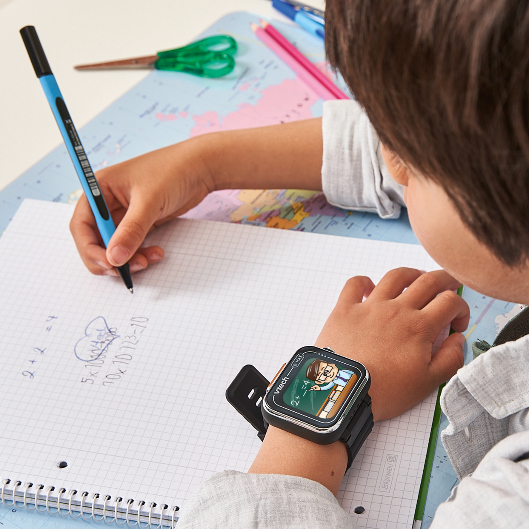 Un niño escribe problemas matemáticos en papel. Un smartwatch muestra un profesor. Tijeras, lápices y un mapa del mundo son visibles.