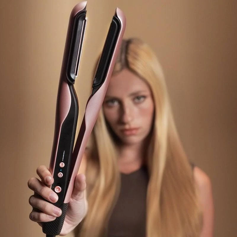 Una mujer sostiene una plancha de pelo rosa y negra. Tiene el pelo largo y rubio y lleva una camiseta oscura.