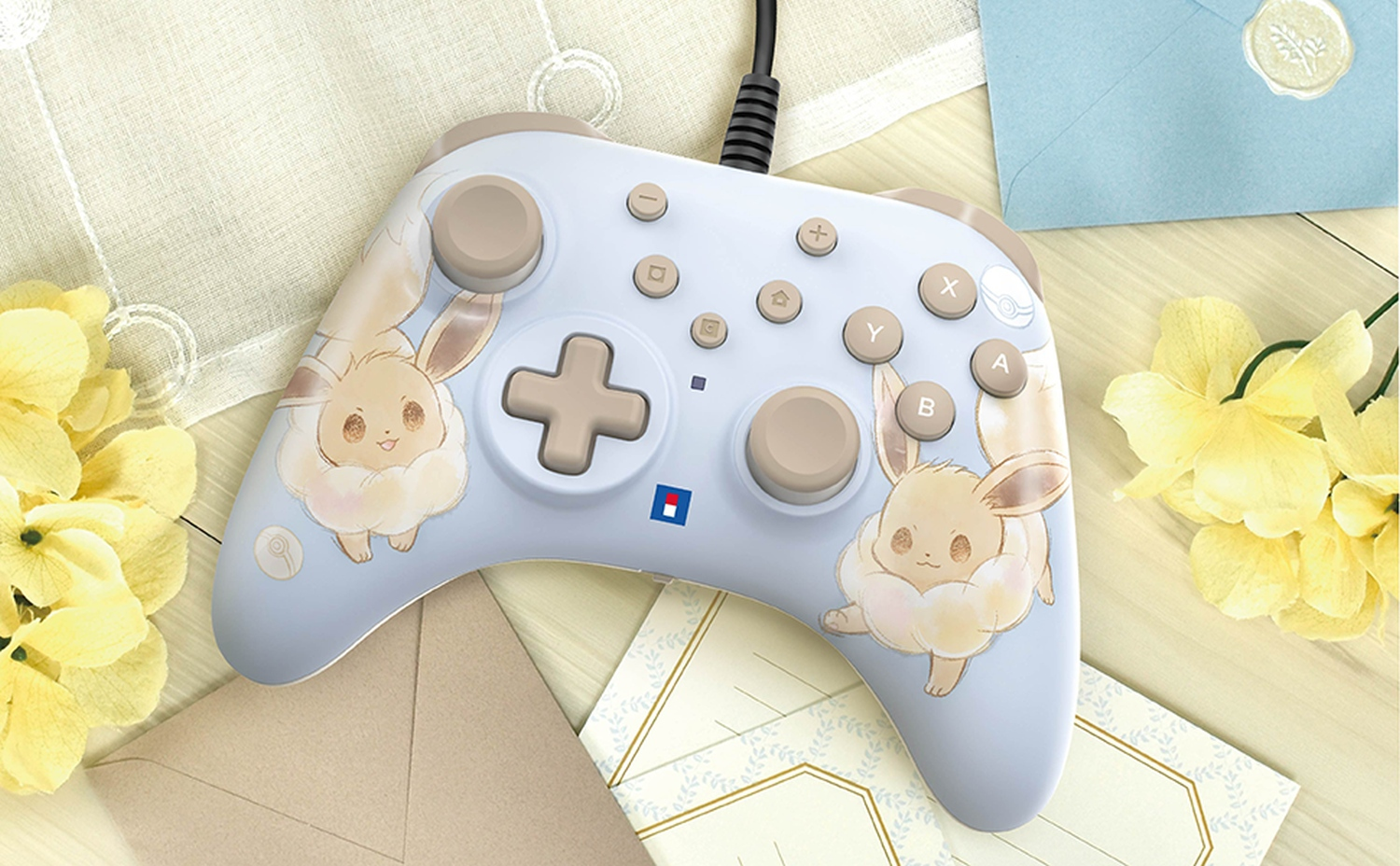 Une manette de jeu bleu clair avec des motifs Évoli, des boutons et un cordon noir. À côté, il y a des fleurs jaunes et des enveloppes.